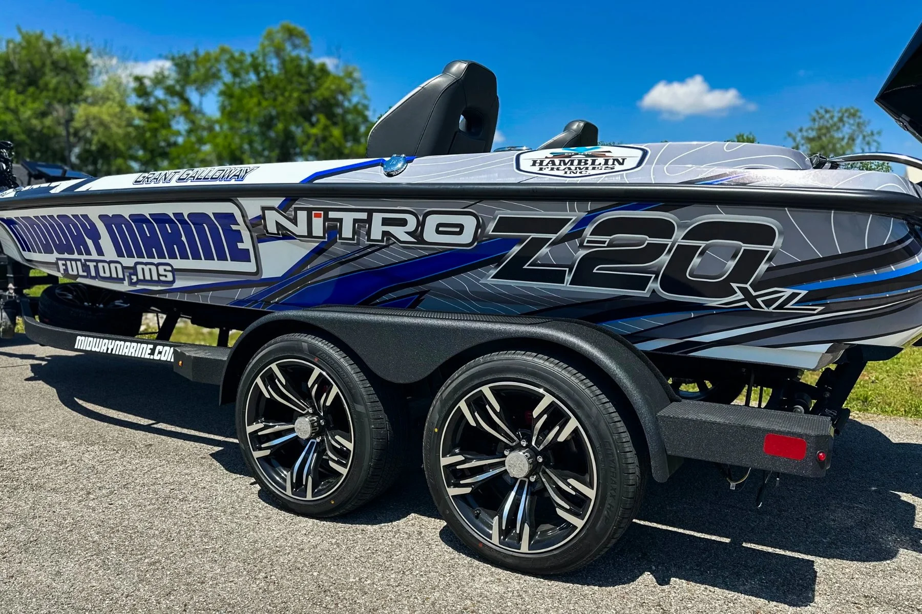 A bass boat on a trailer with a blue, black, grey, and white design, labeled 'Nitro Z20 XL,' parked outdoors on a paved area with green trees and blue sky in the background.
