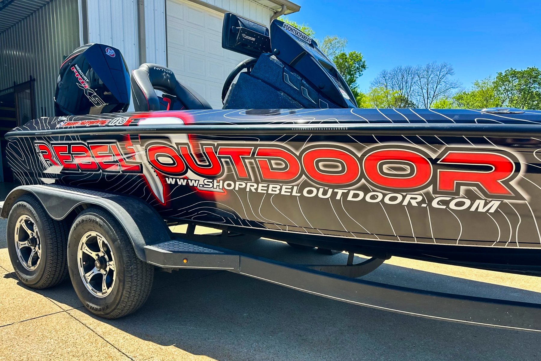 Speedboat with advertising decal reading 'Rebel Outdoor' on side, parked on trailer with tires visible, against a blue sky and trees in background.