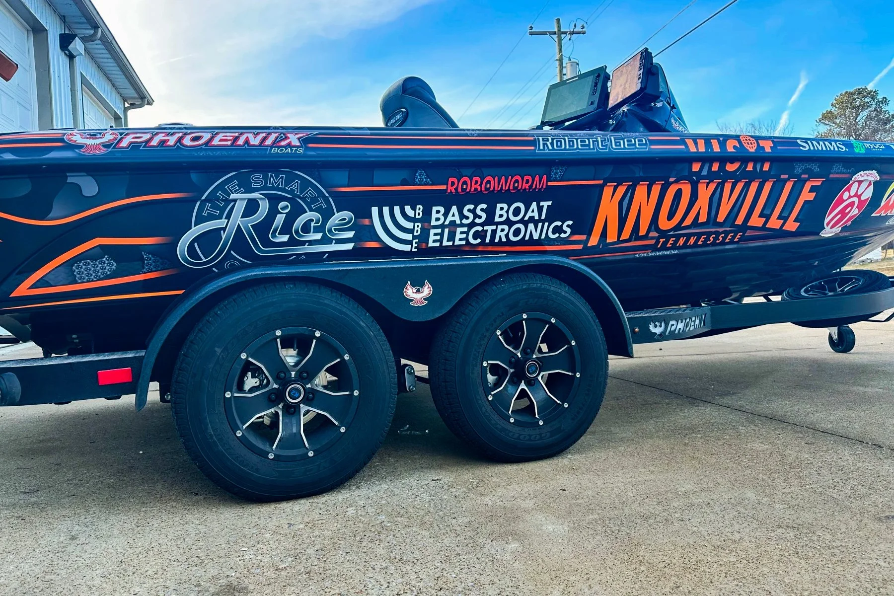 A black boat with multiple sponsor logos and decals, mounted on a trailer, parked on a concrete surface under a clear sky.