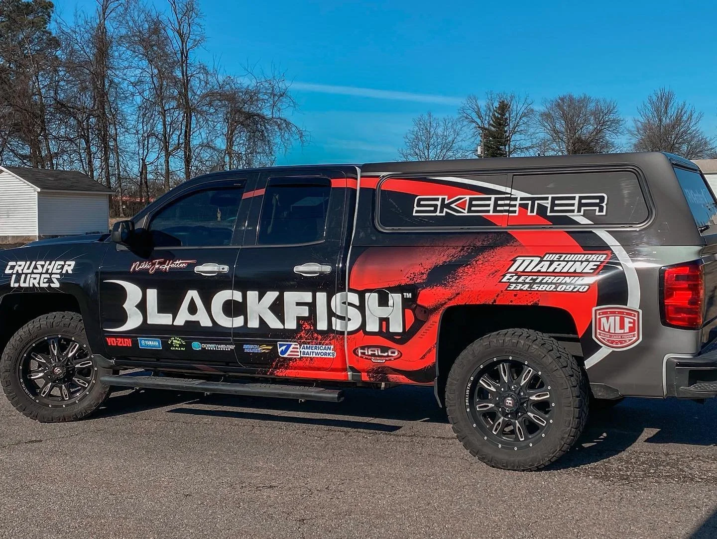 A black pickup truck with various sponsorship and branding graphics, including the large word 'BLACKFISH' on the side, parked outdoors on a paved surface with trees and a house in the background.