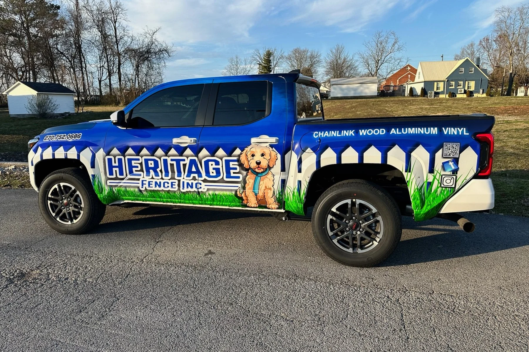 Blue pickup truck with colorful graphics advertising Heritage Fence Inc., featuring a cartoon dog, grass design, and contact information.