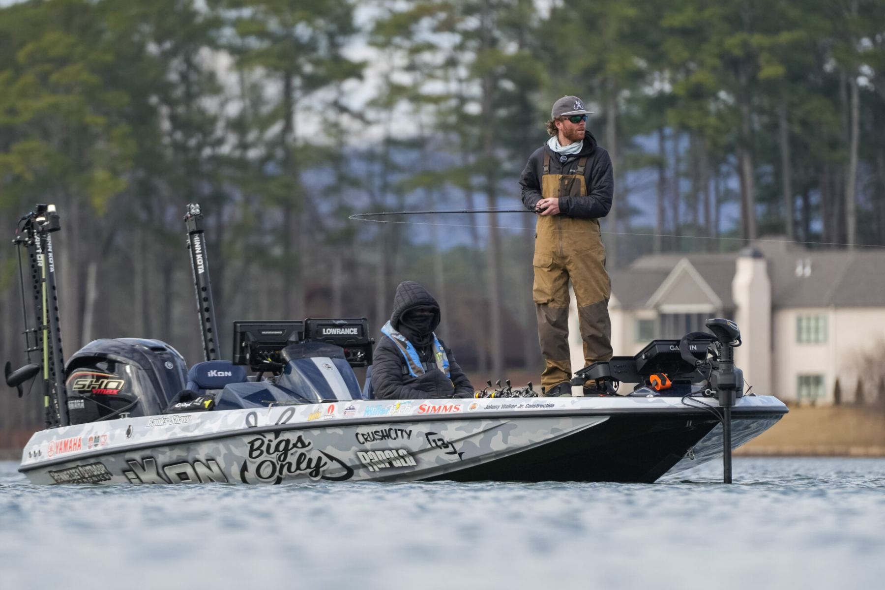 Two men fishing on a boat in a calm lake with houses and trees in the background.