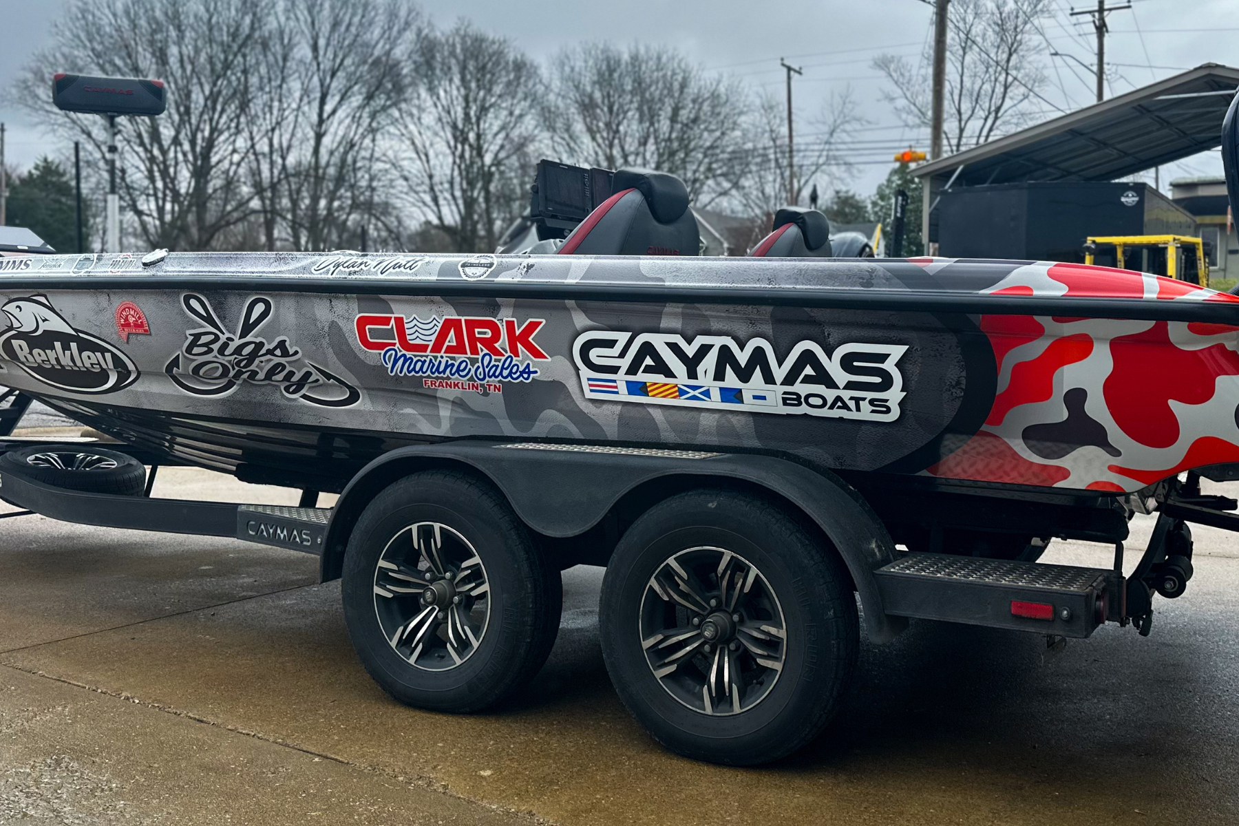 A fishing boat on a trailer with various sponsor logos and a camouflage design, parked on a wet concrete surface, with trees and utility poles in the background.