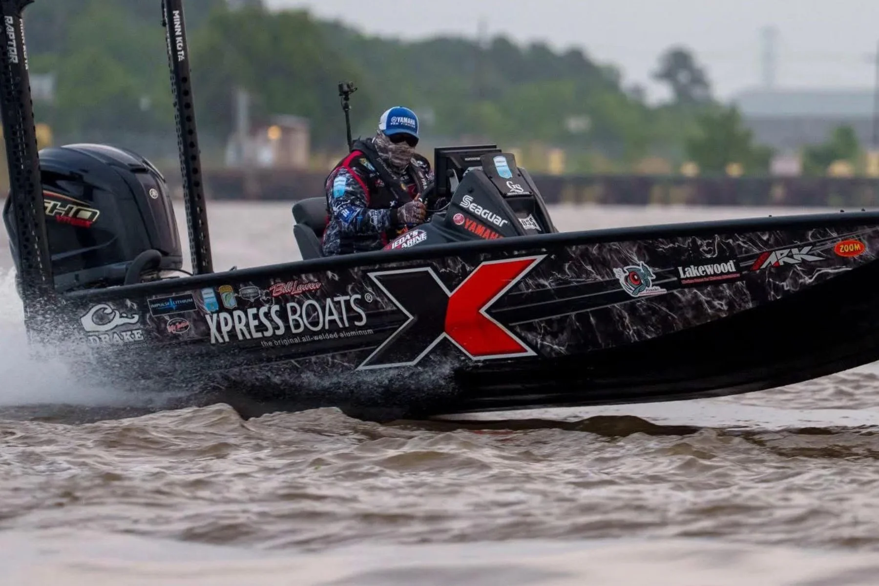 A fisherman on a black aluminum boat with red and white decals, wearing a blue cap and a jacket, navigating the river.