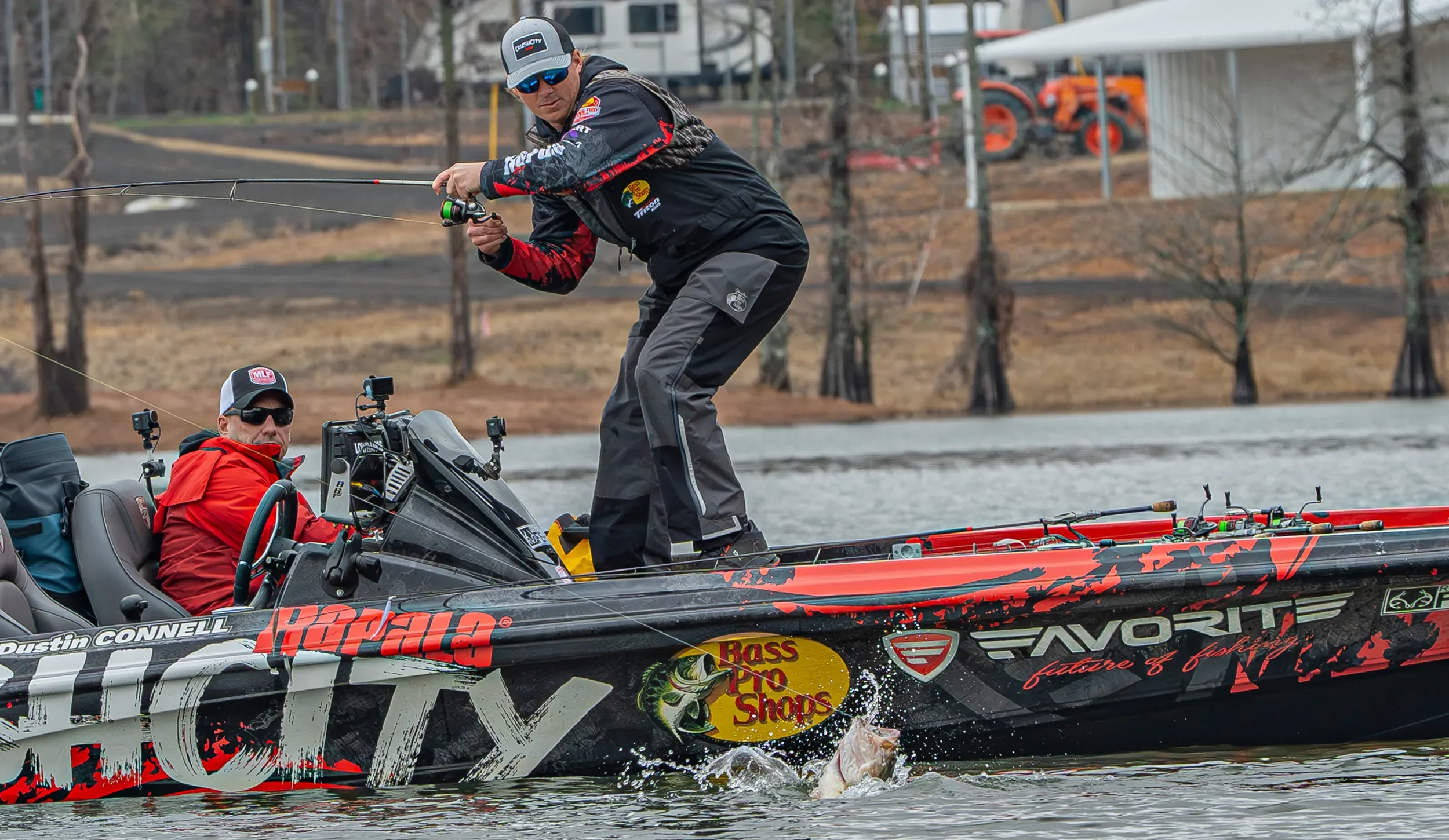 Two fishermen on a boat catching a fish in a lake, with one fisherman standing and reeling in the fish while the other man sits and looks on.