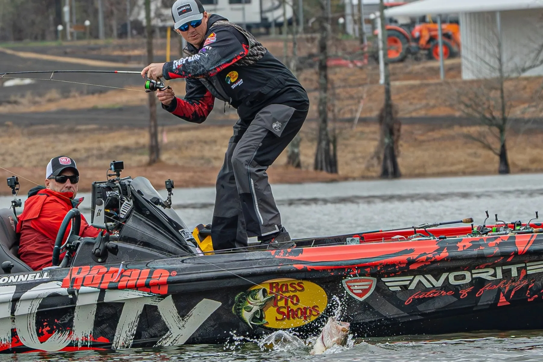 Two men on a fishing boat, one standing and fishing, the other sitting and guiding the boat. The boat is decorated with logos including Bass Pro Shops, Procraft, Atlantic, and Rapala. The man standing is wearing a black jacket, gray pants, sunglasses