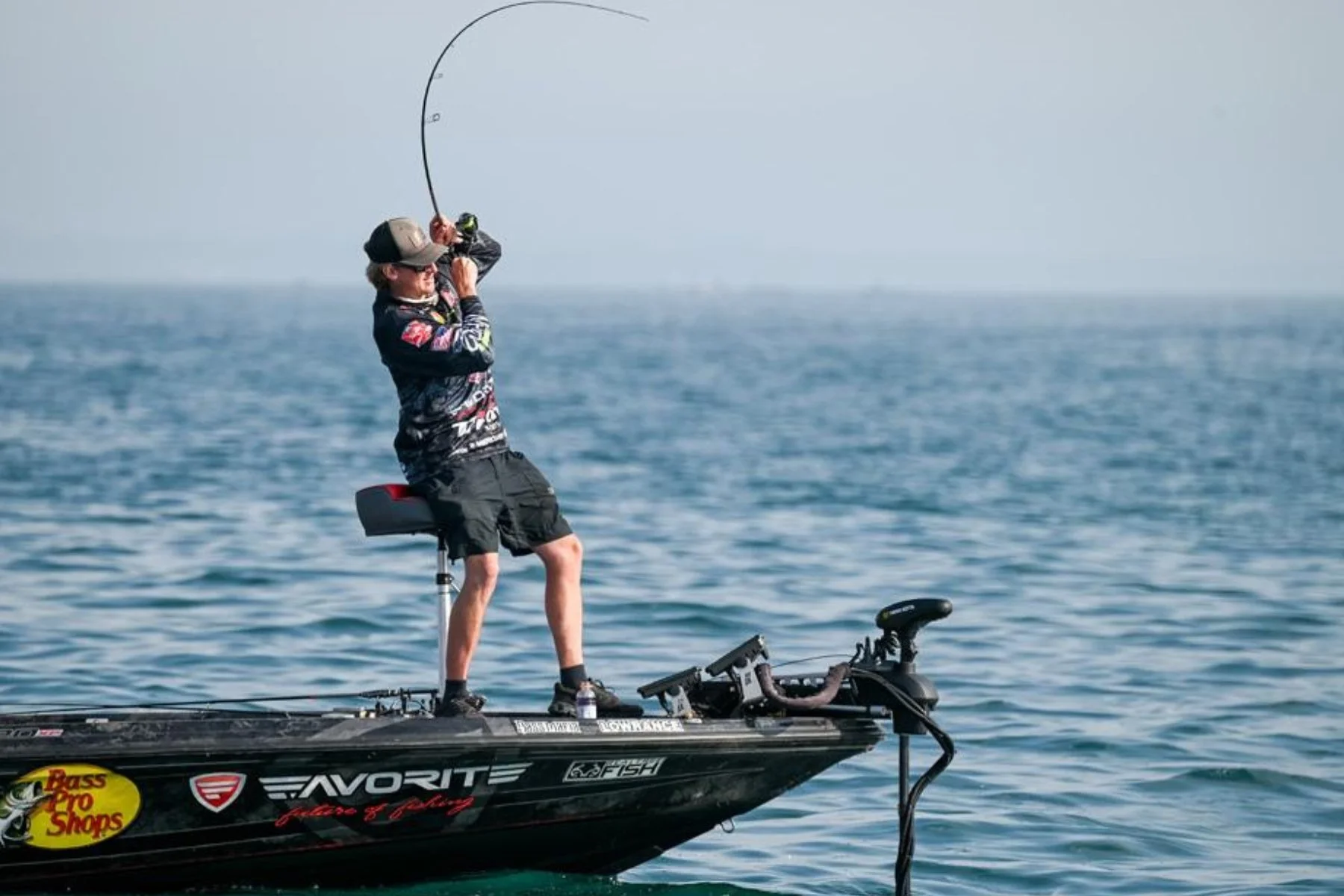 A man fishing on a boat out on the ocean, wearing a cap, sunglasses, and fishing gear, holding a fishing rod with a fish hook in the water.