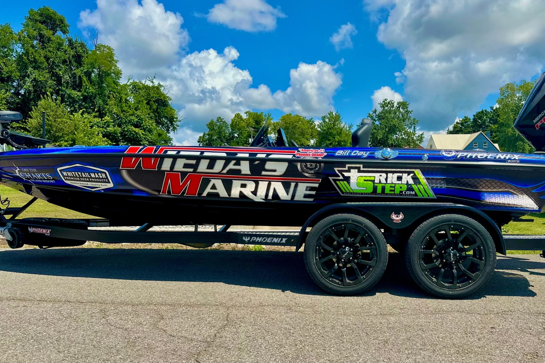 A speedboat on a trailer with various sponsor logos, parked on a paved surface with trees and a cloudy sky in the background.