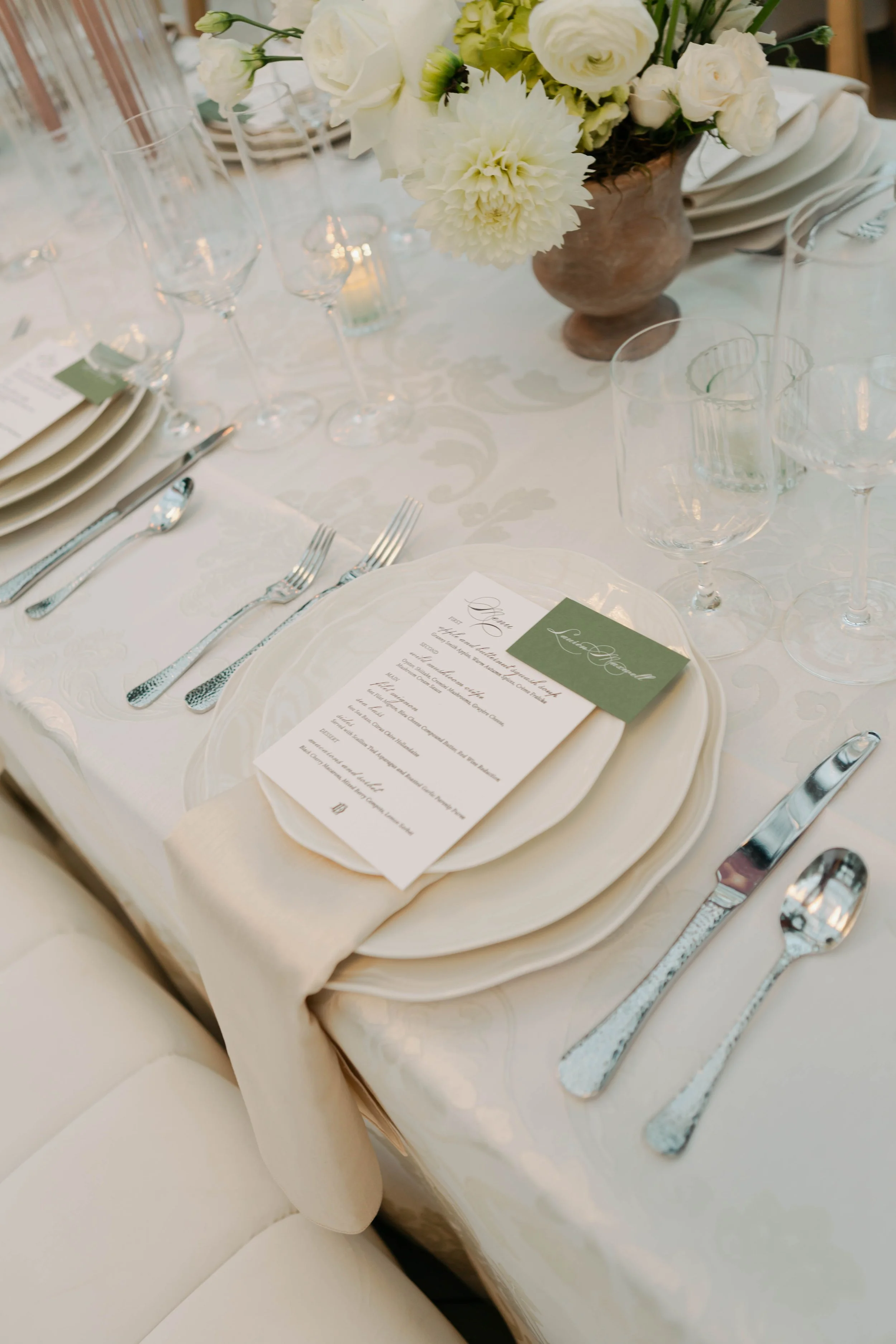 Elegant table setting with white plates, silverware, and a menu card on a white tablecloth, decorated with a bouquet of white flowers in a brown vase.