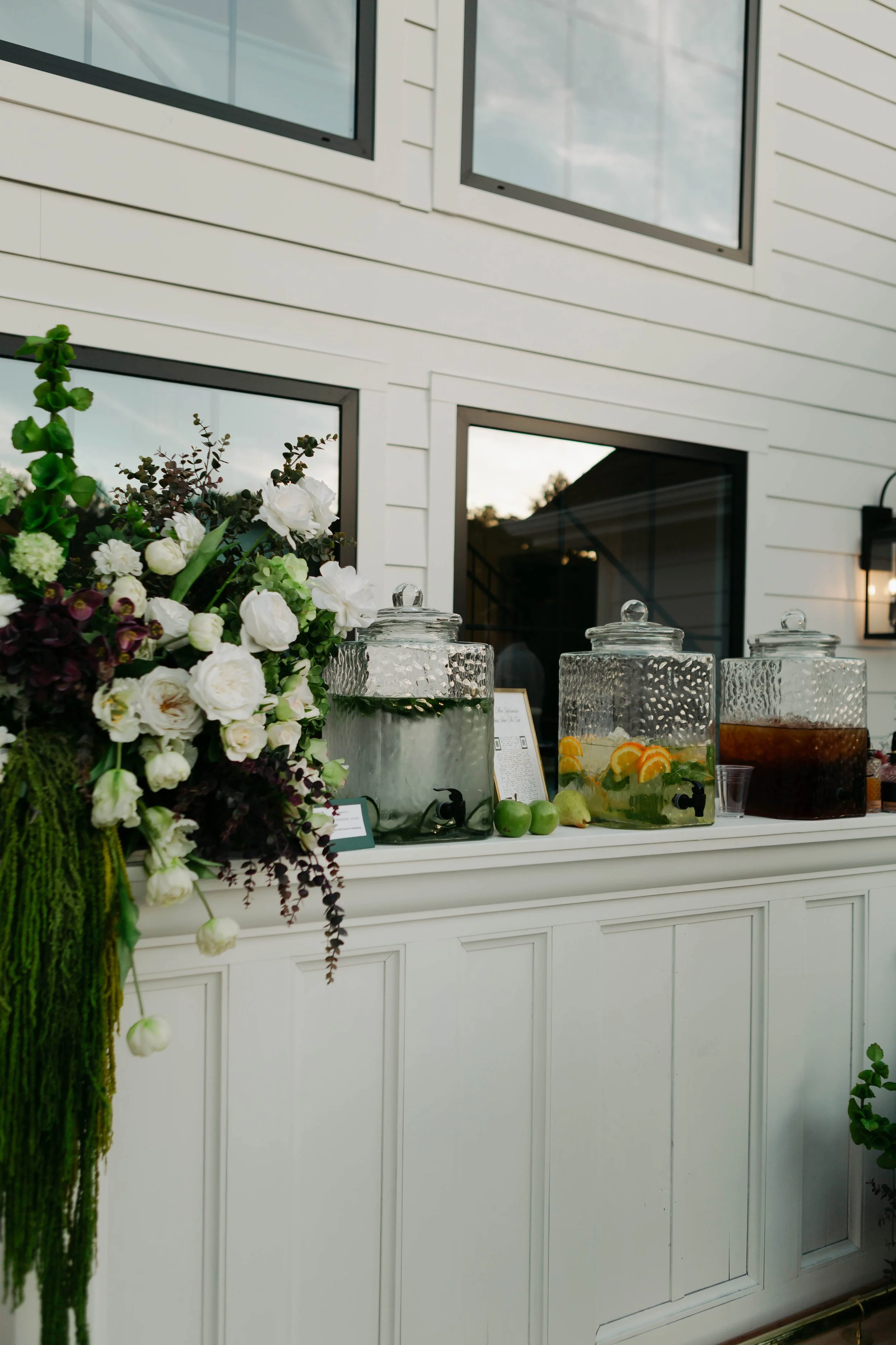 Indoor or outdoor setting with large glass beverage dispensers filled with water and fruit slices, arranged on a white counter next to a floral arrangement with white and dark purple flowers.