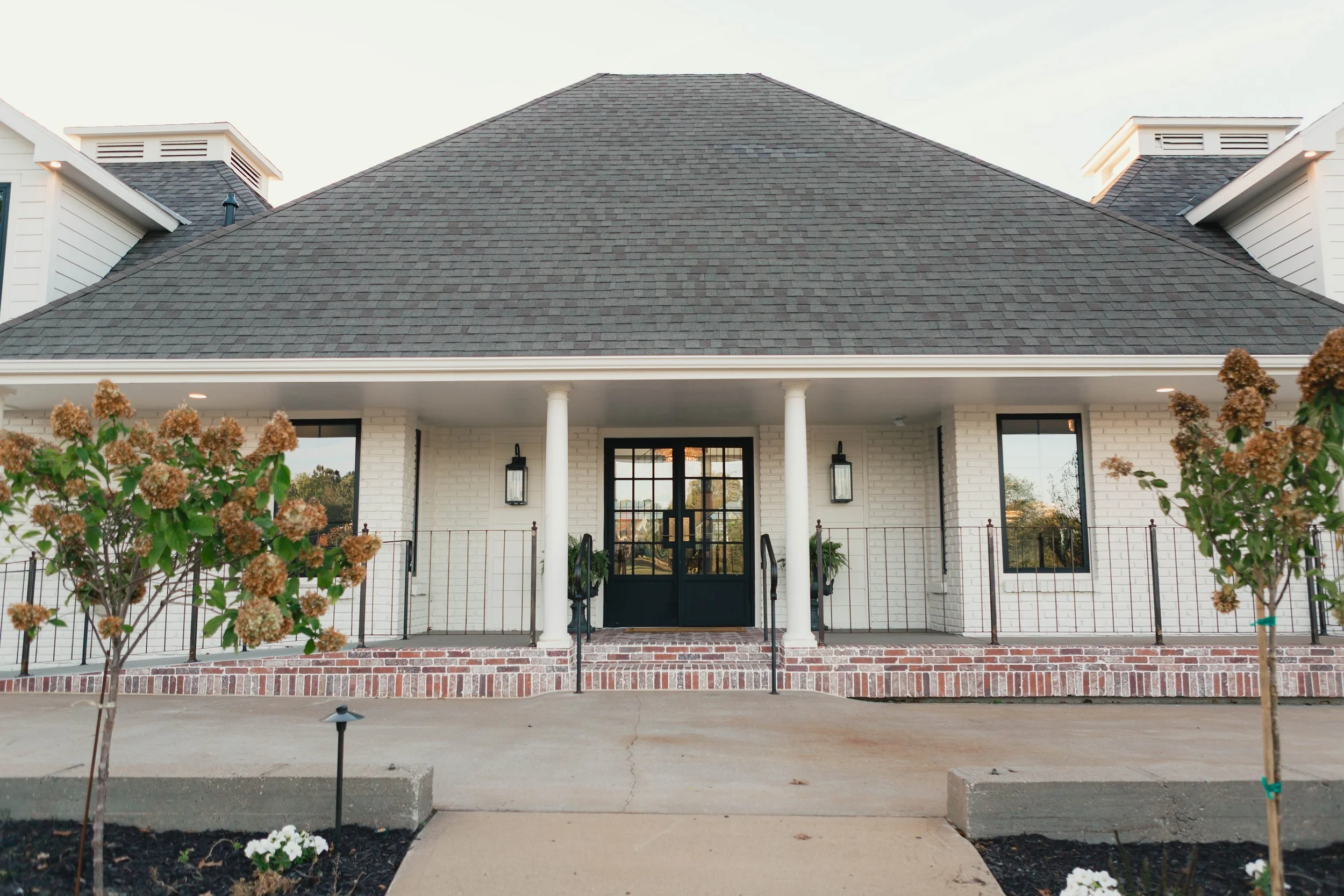 Front view of a white brick house with a dark gray shingled roof, entrance with double glass doors, and a porch with white columns and black railings.