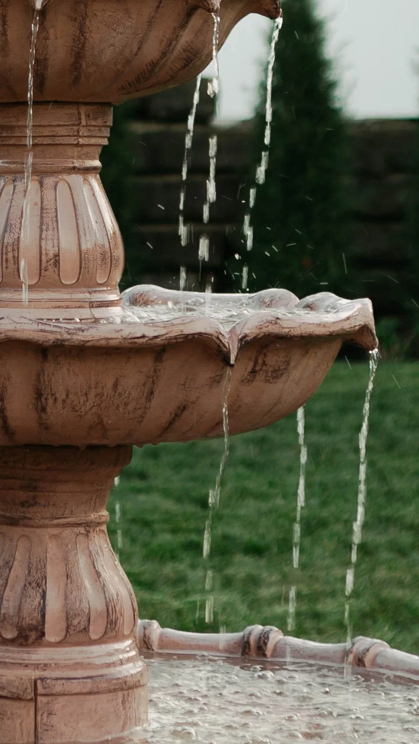 Close-up of a tiered outdoor fountain with water flowing from the top tier to the bottom, set in a garden with green grass and blurred trees in the background.