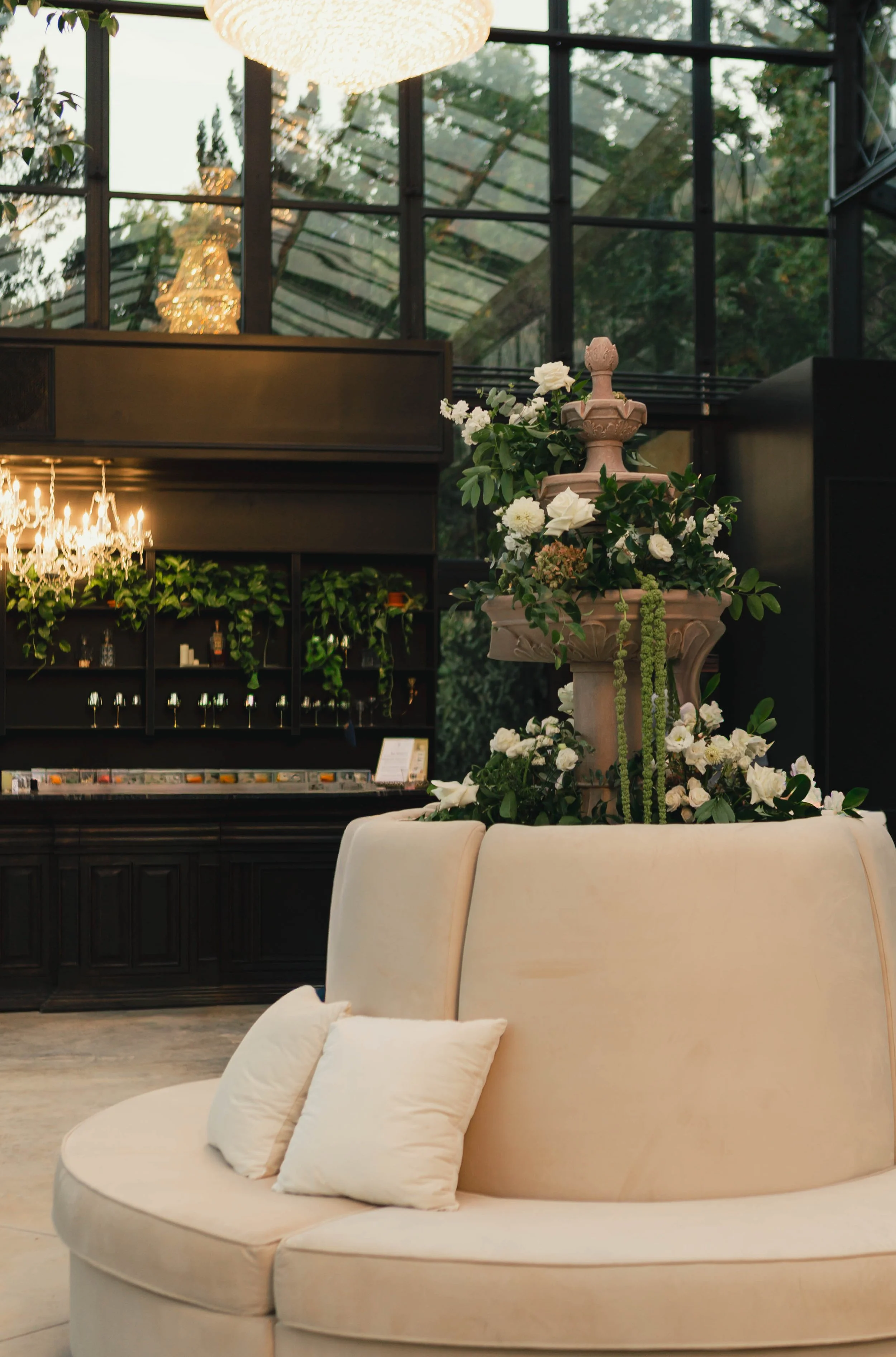 Interior of a luxury lounge with a curved cream sofa, white pillows, a floral arrangement, a large chandelier, dark wood shelves, glassware, and greenery in the background.