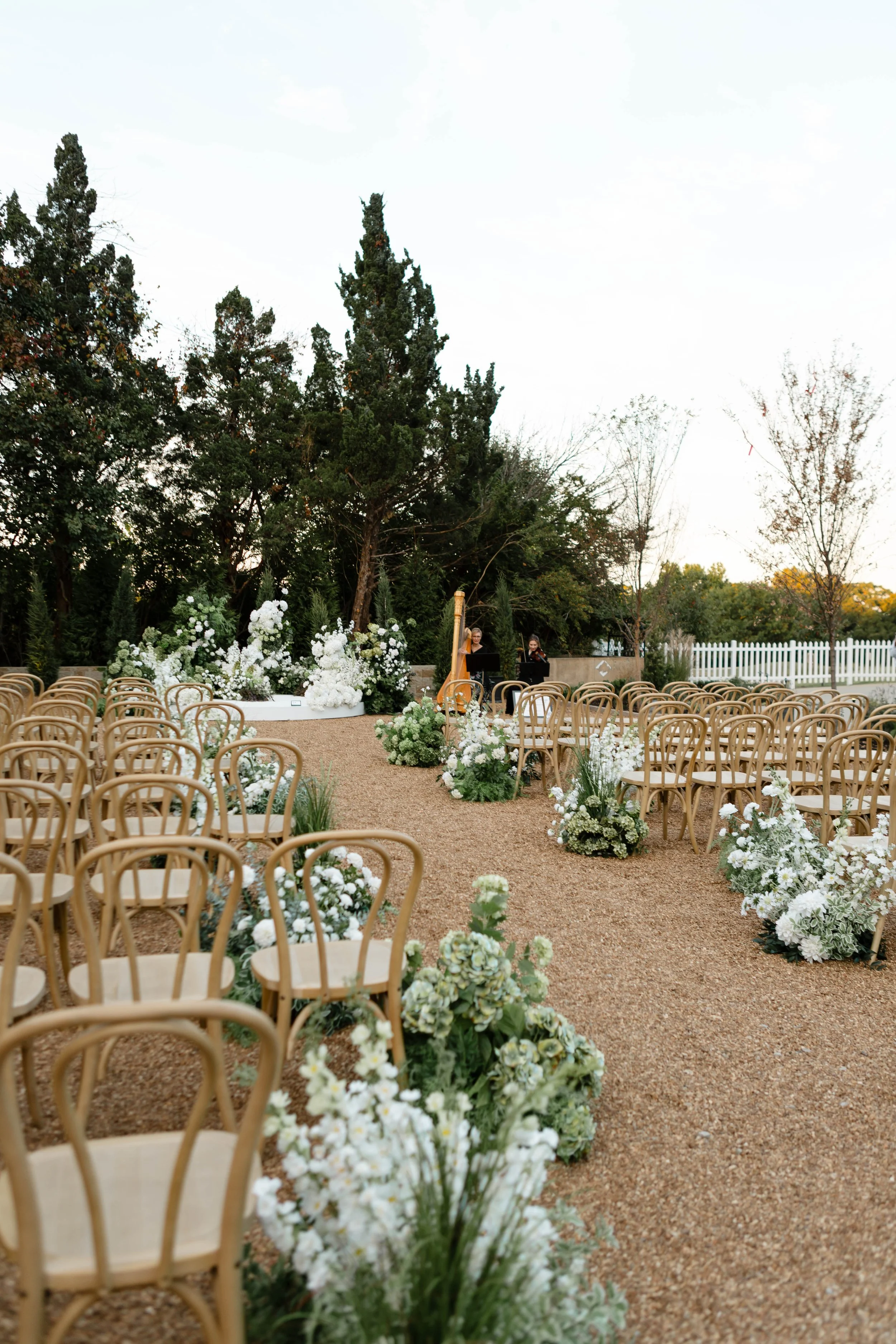Outdoor wedding ceremony setup with rows of wooden chairs, white floral arrangements along the aisle, and a harp player in the background, surrounded by trees.