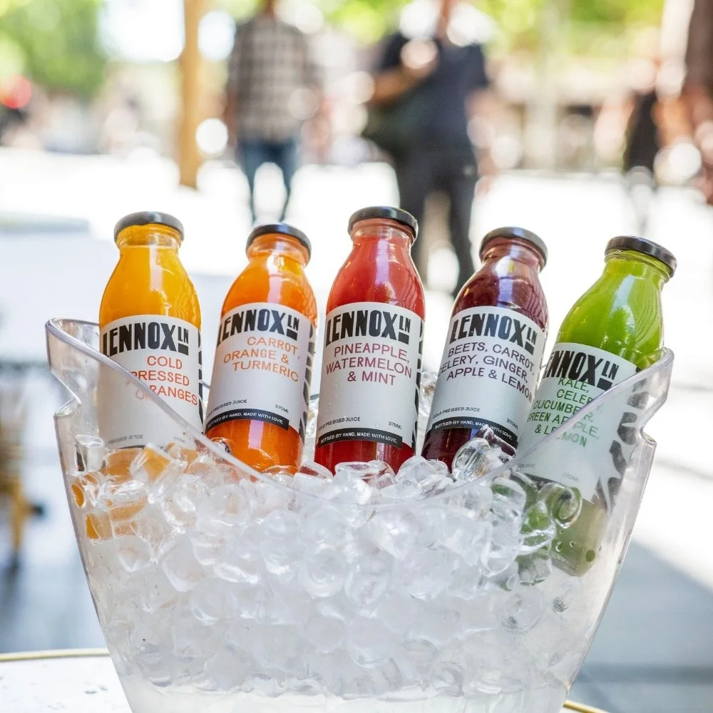 Five bottles of colorful juices in a clear ice bucket outdoors, with people blurred in the background.