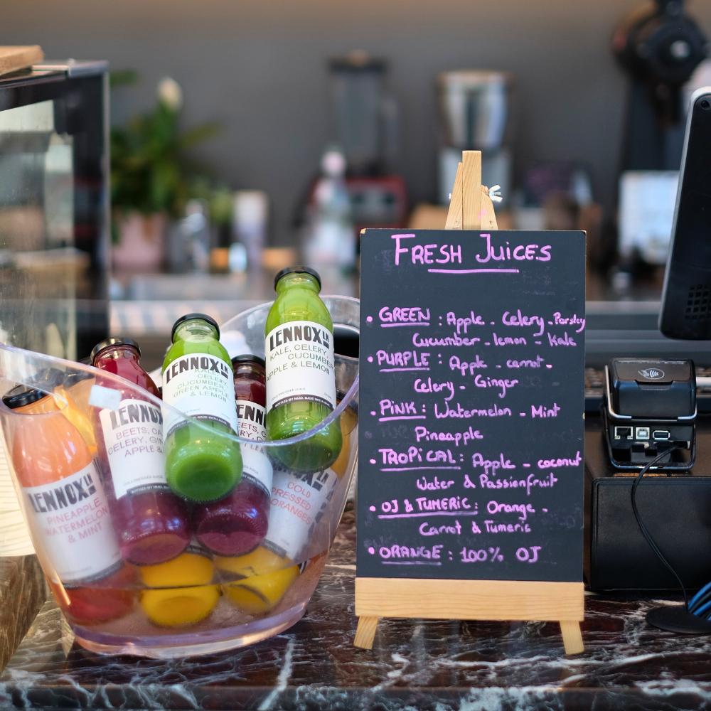 A small chalkboard sign listing different fresh juice options and ingredients, placed next to bottles of colorful juices on a marble countertop. The juice options include green, purple, pink, tropical, OJ & turmeric, and orange.