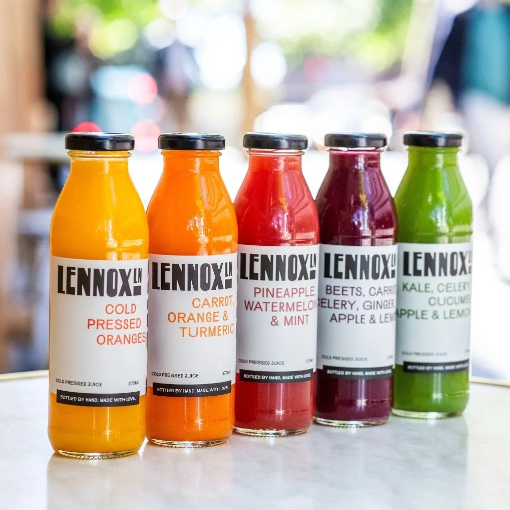Five bottles of cold-pressed juice with black caps, labeled with various flavors and ingredients, arranged in a row on a light-colored surface with a blurred outdoor background.