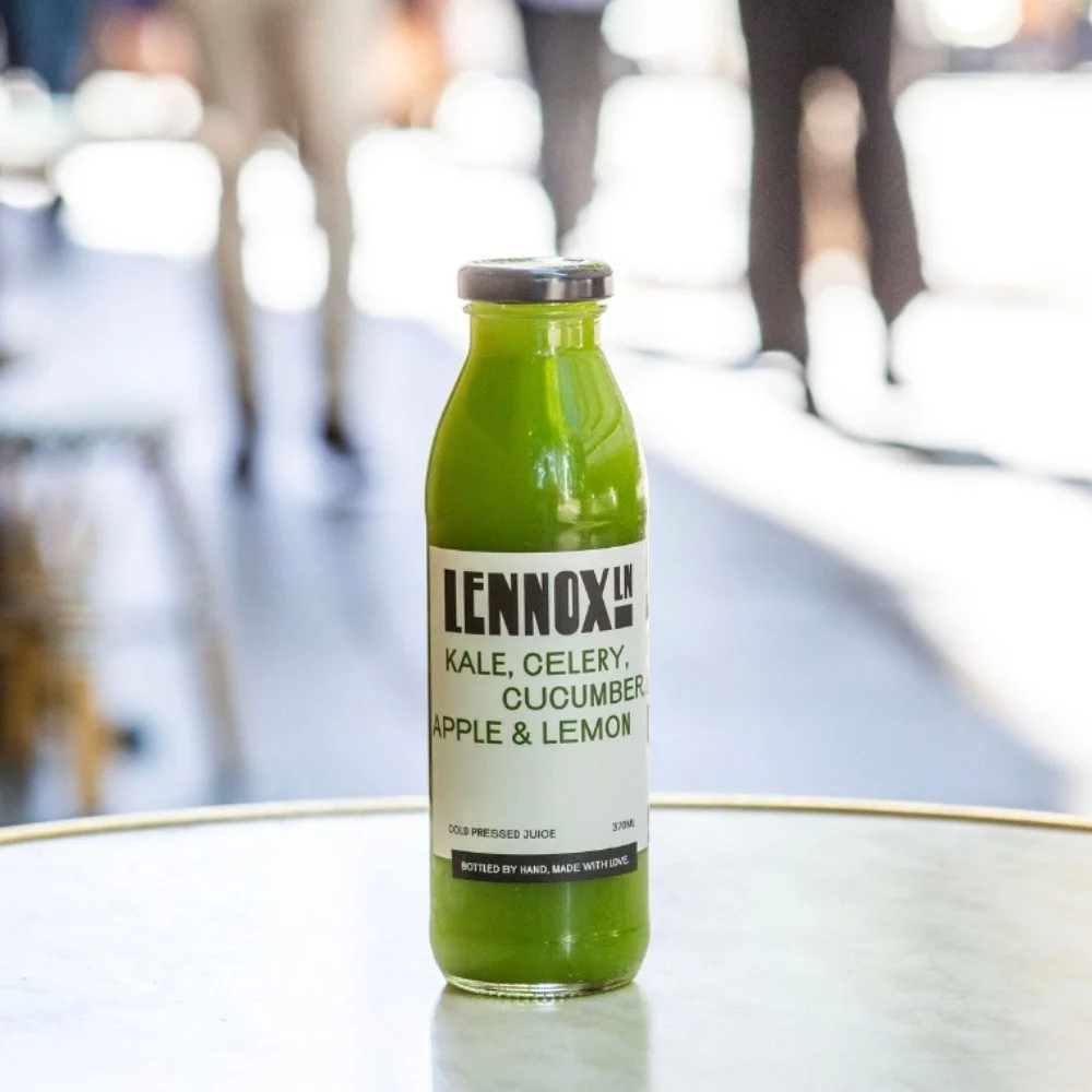 Green bottled juice labeled 'Lennox In' with ingredients kale, celery, cucumber, apple, and lemon, placed on a white surface in a bright, busy setting.