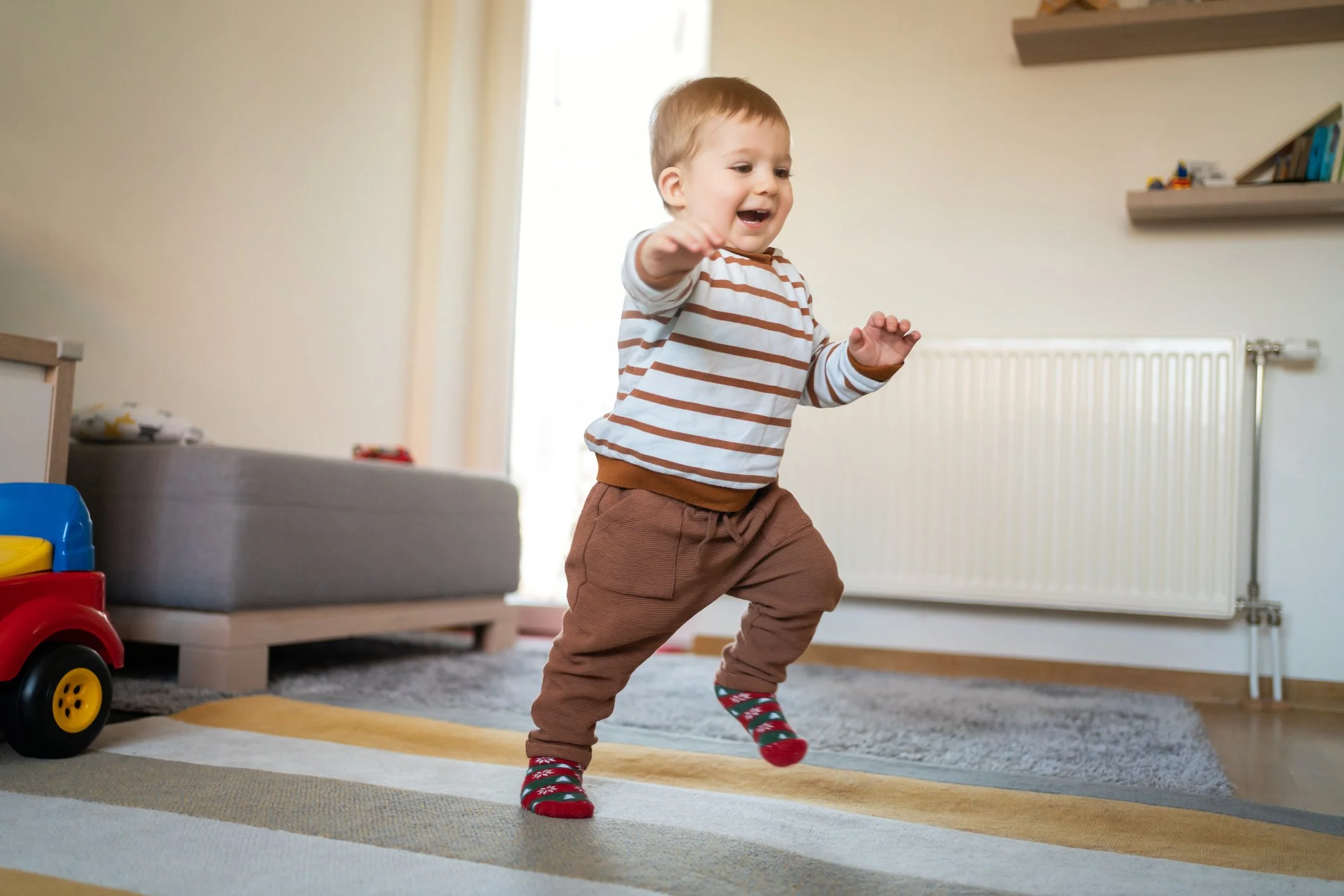 A young boy with short light brown hair wearing a white and brown striped long sleeve shirt, brown pants, and festive red and green socks, smiling and reaching out as he walks across a room with a colorful rug and toys.