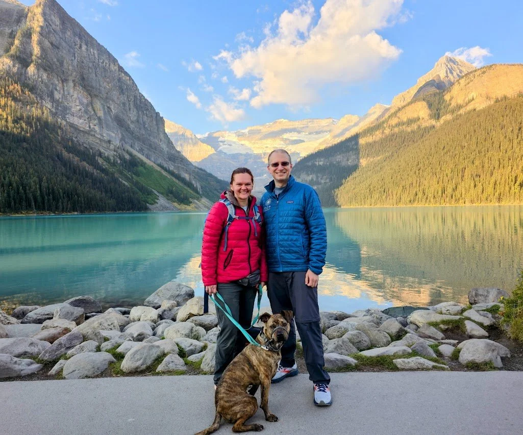 A smiling couple with a dog standing on a rocky shore by a turquoise lake, surrounded by mountains and trees, under a blue sky with clouds.