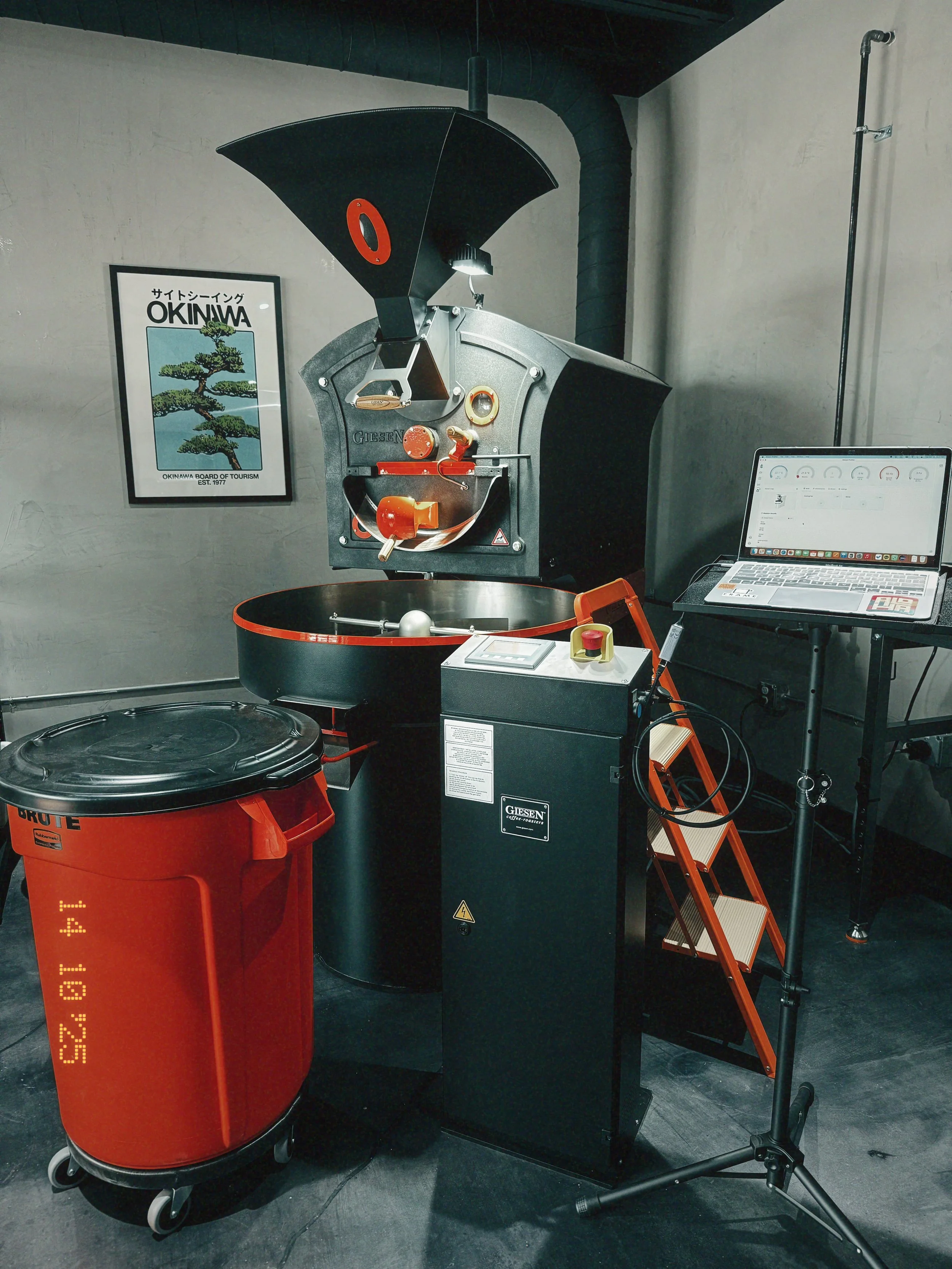 A Giesen coffee roasting machine with a computer monitor next to it, in a room with a framed poster of Okinawa on the wall.