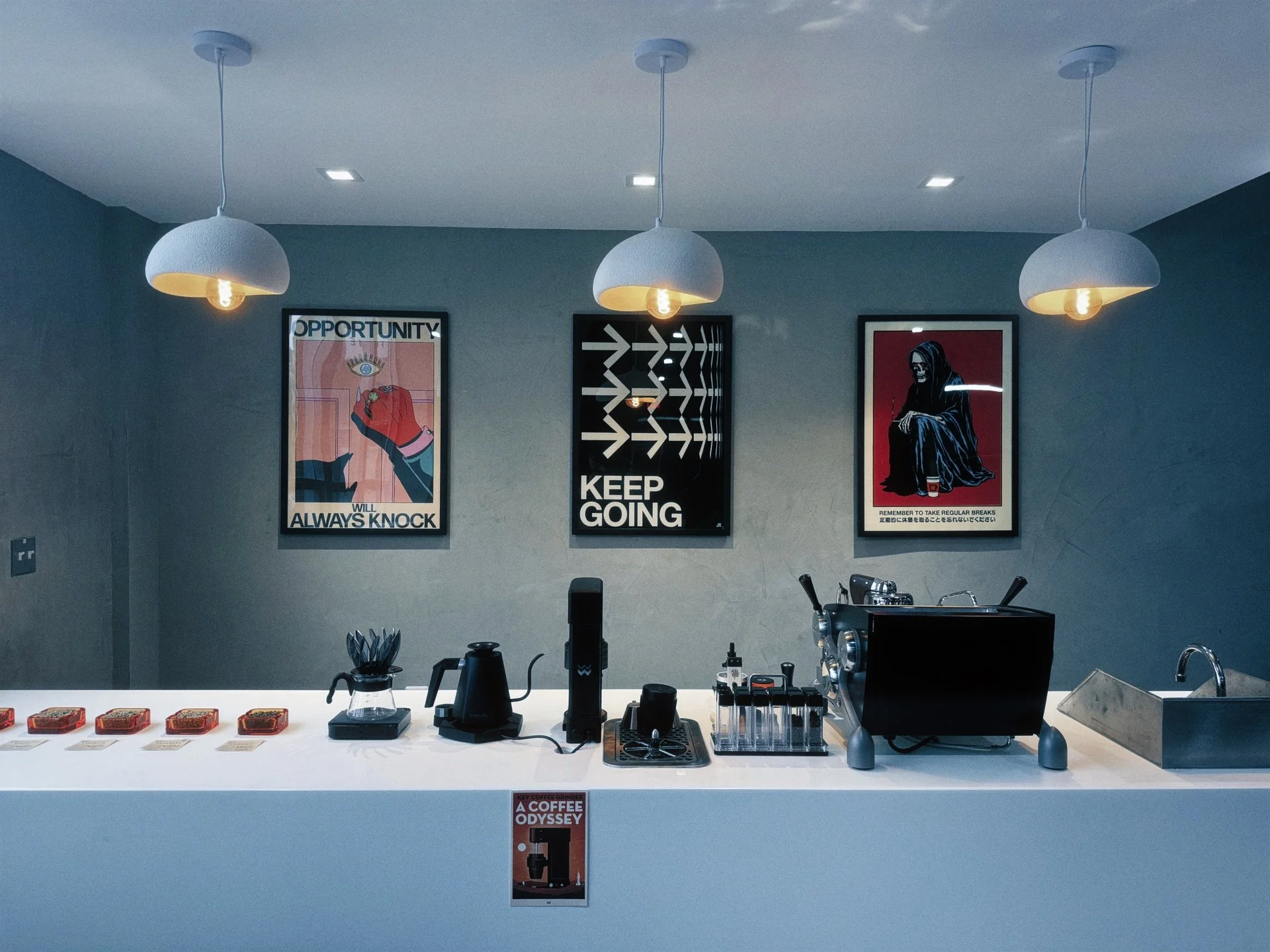 Modern coffee shop counter with three hanging pendant lights, framed motivational posters on the wall, and various coffee-making equipment including a coffee grinder, kettle, test tubes, and a stainless steel sink.