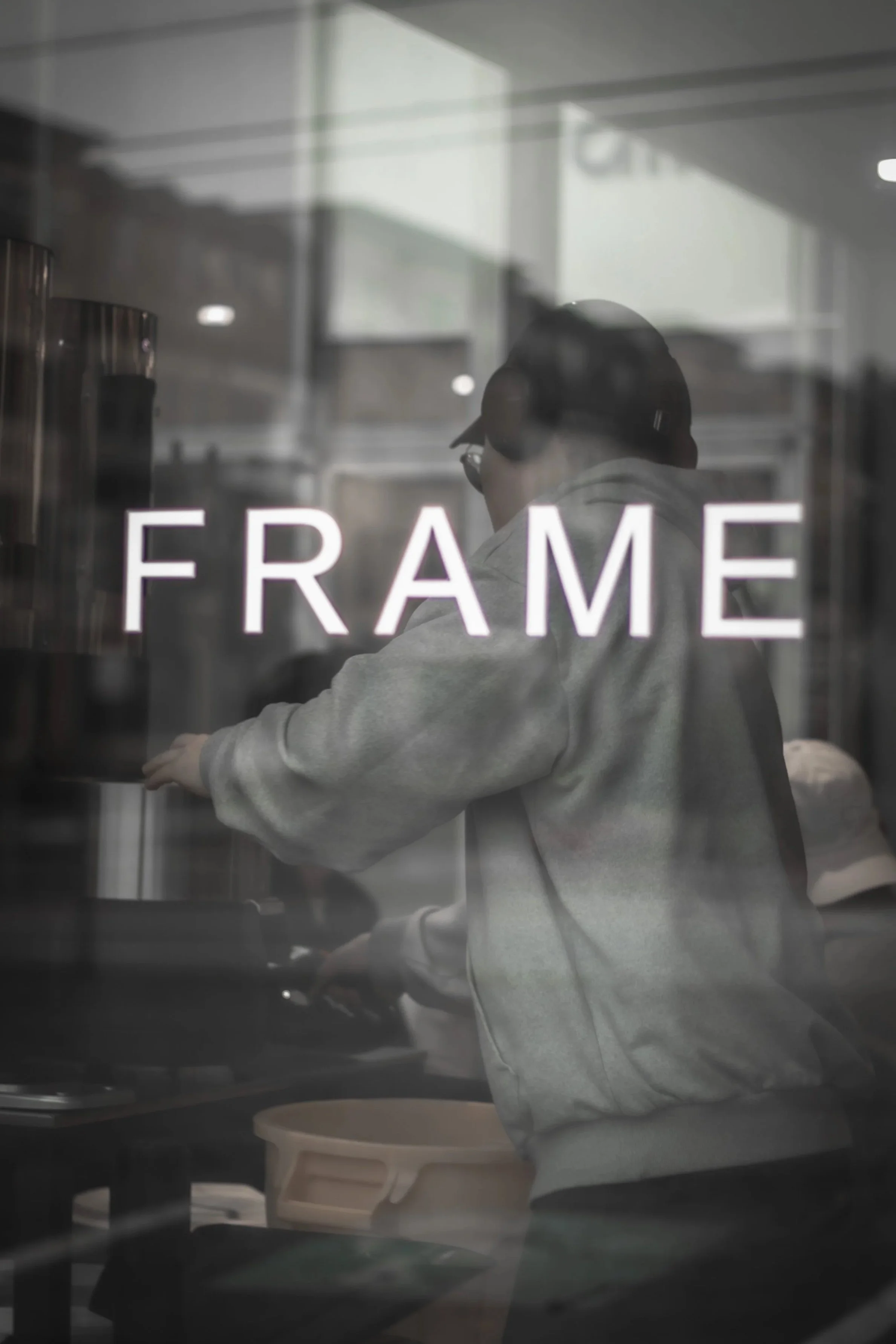 A person with a gray hoodie and hat seen through a glass wall with the word 'FRAME' written on it, inside what appears to be a kitchen or food establishment.