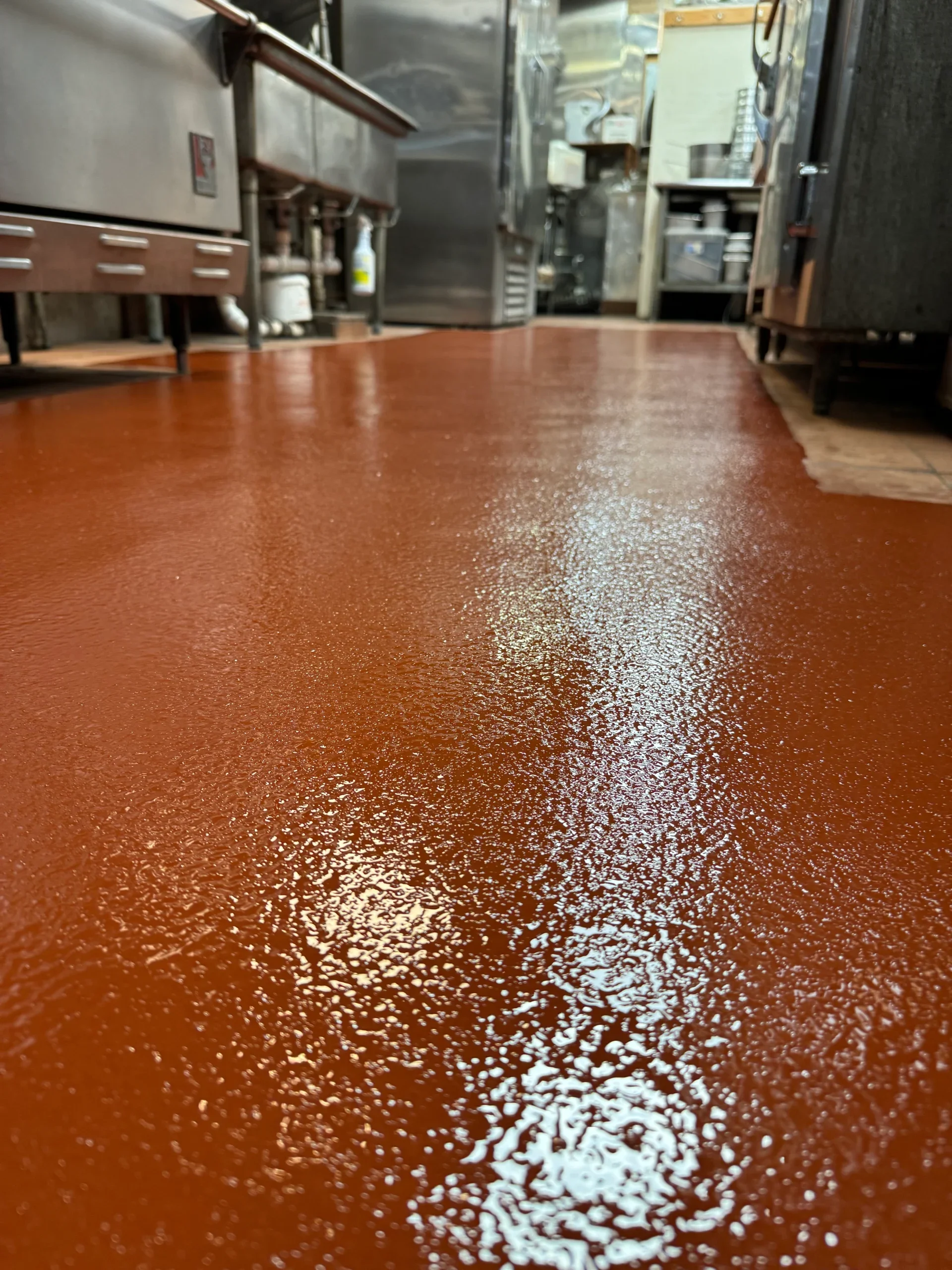 A view of a freshly coated, shiny reddish-brown floor in a commercial kitchen with stainless steel appliances and equipment.