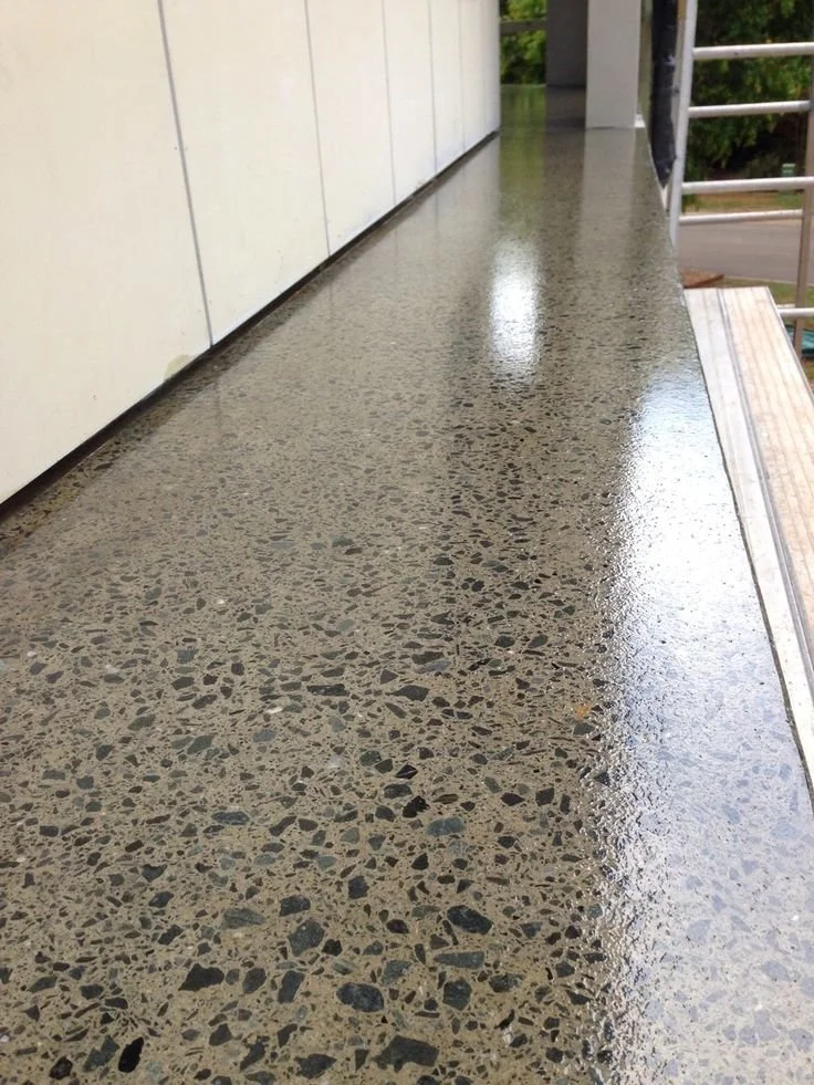 Close-up of a wet concrete balcony or porch with a textured surface, next to a white wall and metal railing, outdoors.