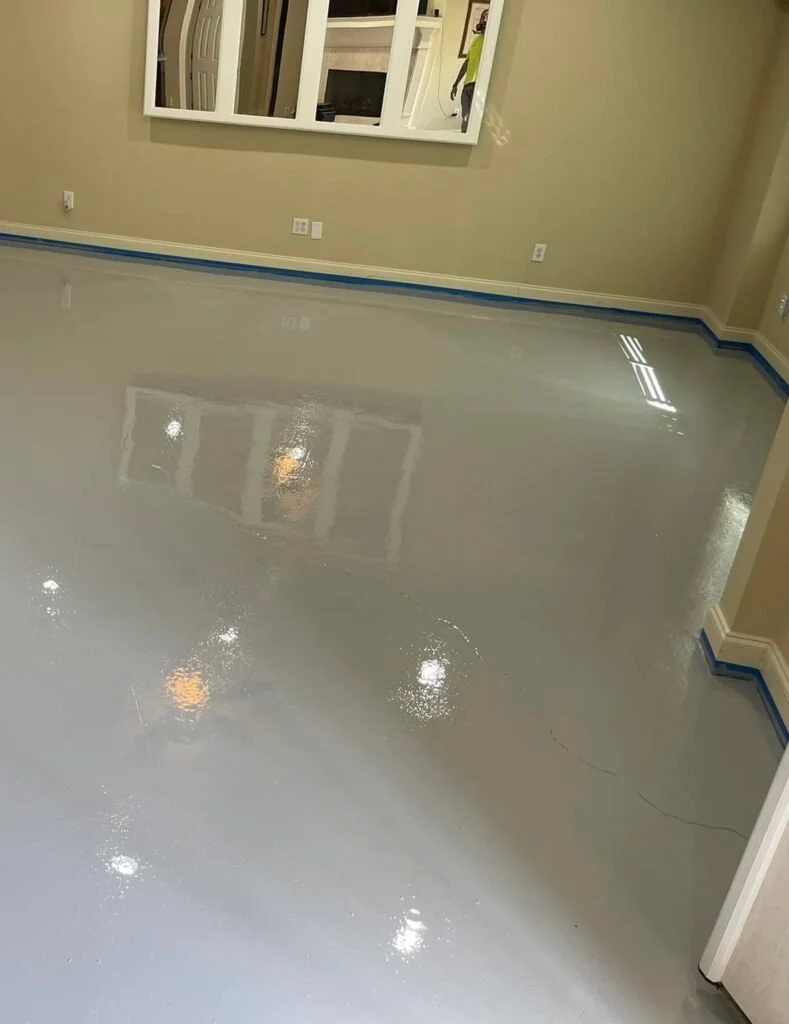Empty room with newly poured light gray epoxy floor, blue painter's tape along the edges, and a large mirror reflecting a fireplace and a person in a yellow shirt.
