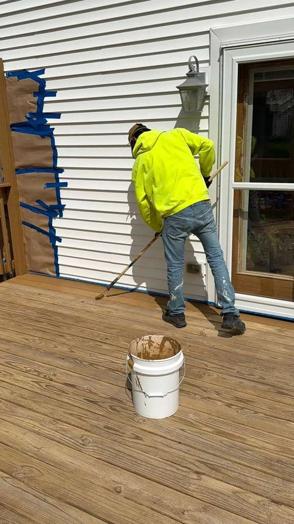 A person in a yellow hoodie and jeans is staining a wooden deck with brown stain using a long-handled brush. There is a bucket of stain on the deck, and the person is painting near the corner of a white house with a sliding glass door.