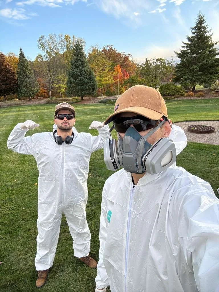 Two men dressed in white protective suits and safety gear, standing outdoors on well-manicured grass with trees showing fall foliage in the background. One man in the foreground is wearing a cap, sunglasses, and a respirator mask, while the other is