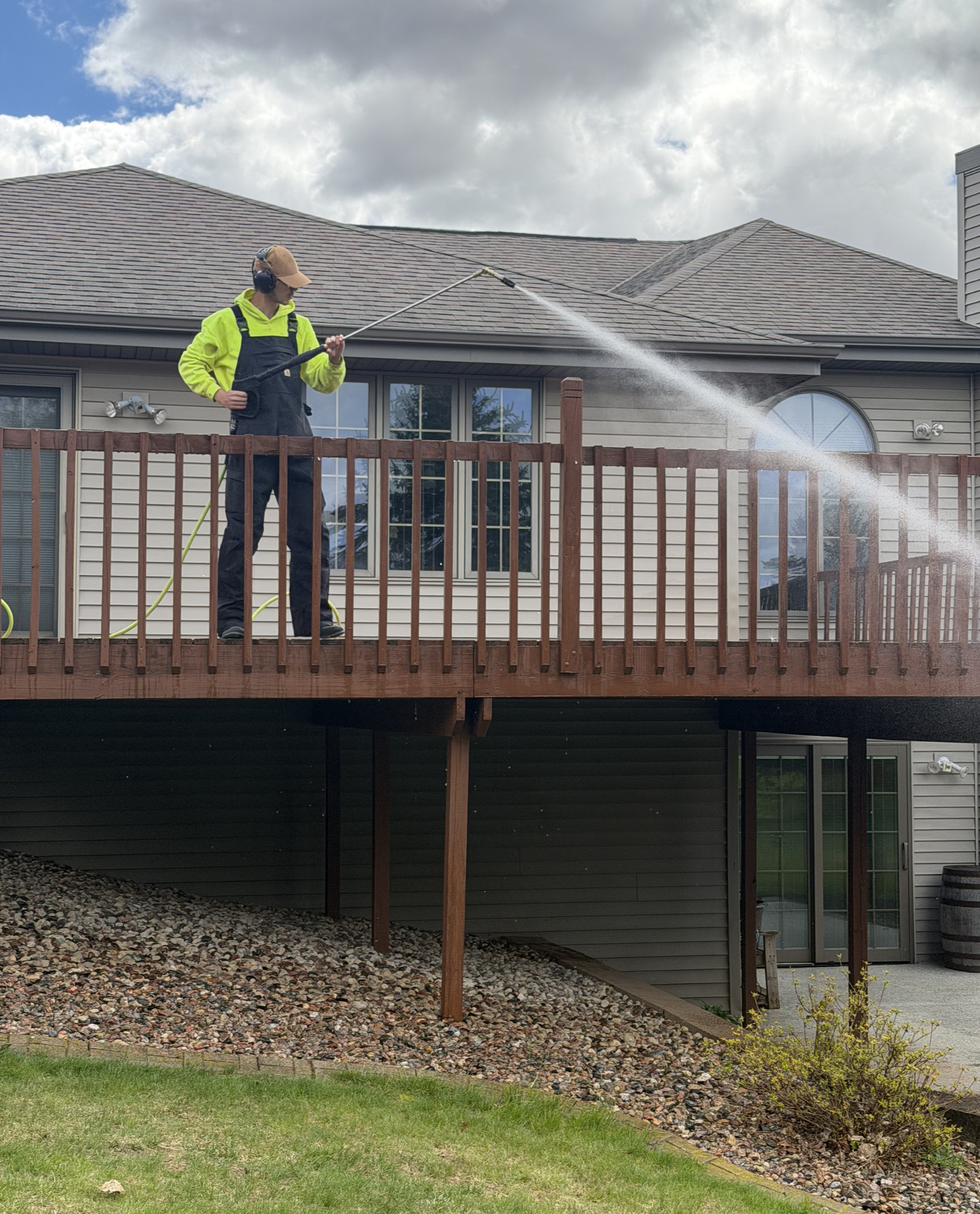 Person wearing a yellow hoodie and black overalls pressure washing the roof of a house from a wooden deck.