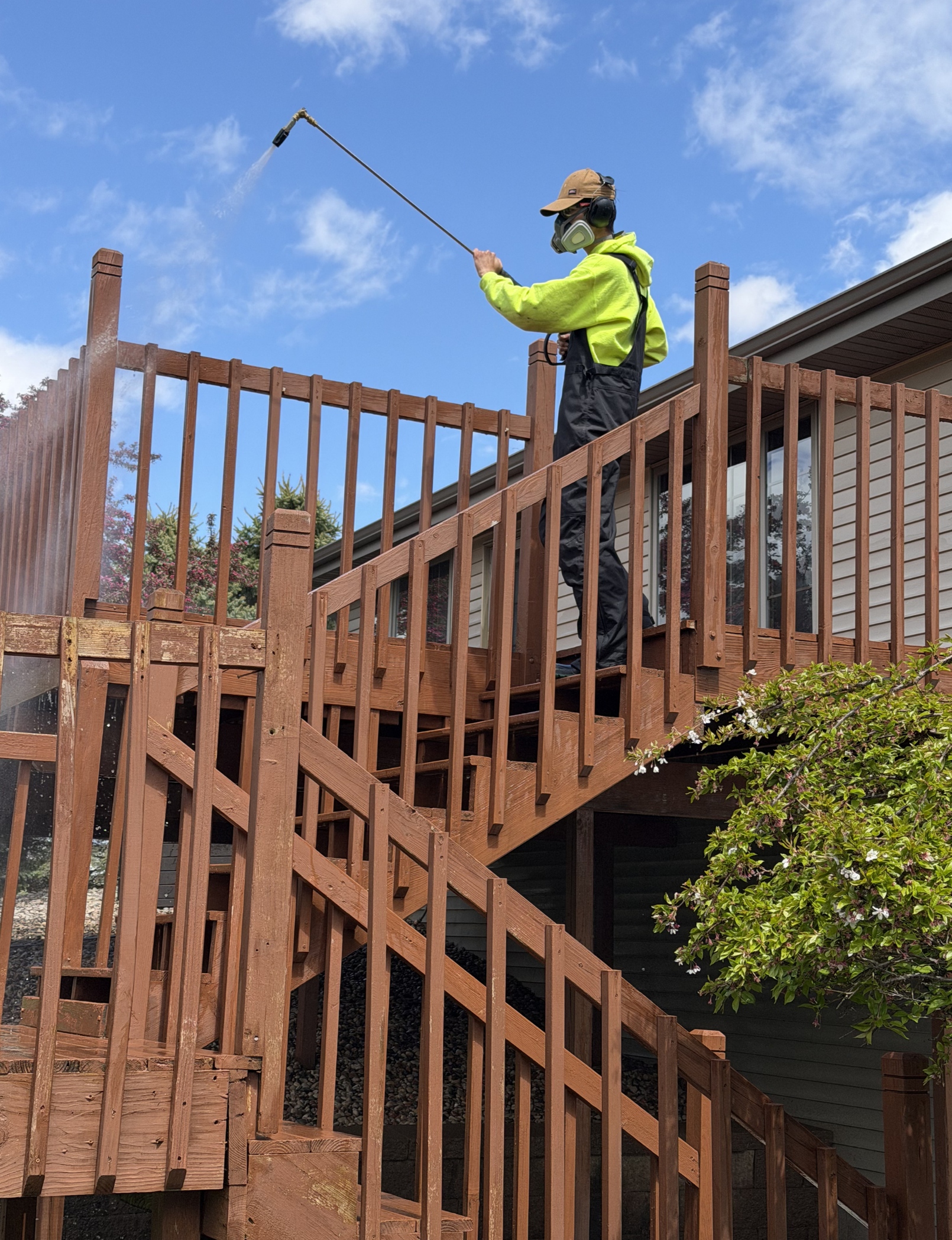 A person in a yellow hoodie and black pants cleaning a wooden staircase railing with a power washer, wearing a hat, safety glasses, and ear protection, outside a house with a cloudy sky and some trees in the background.