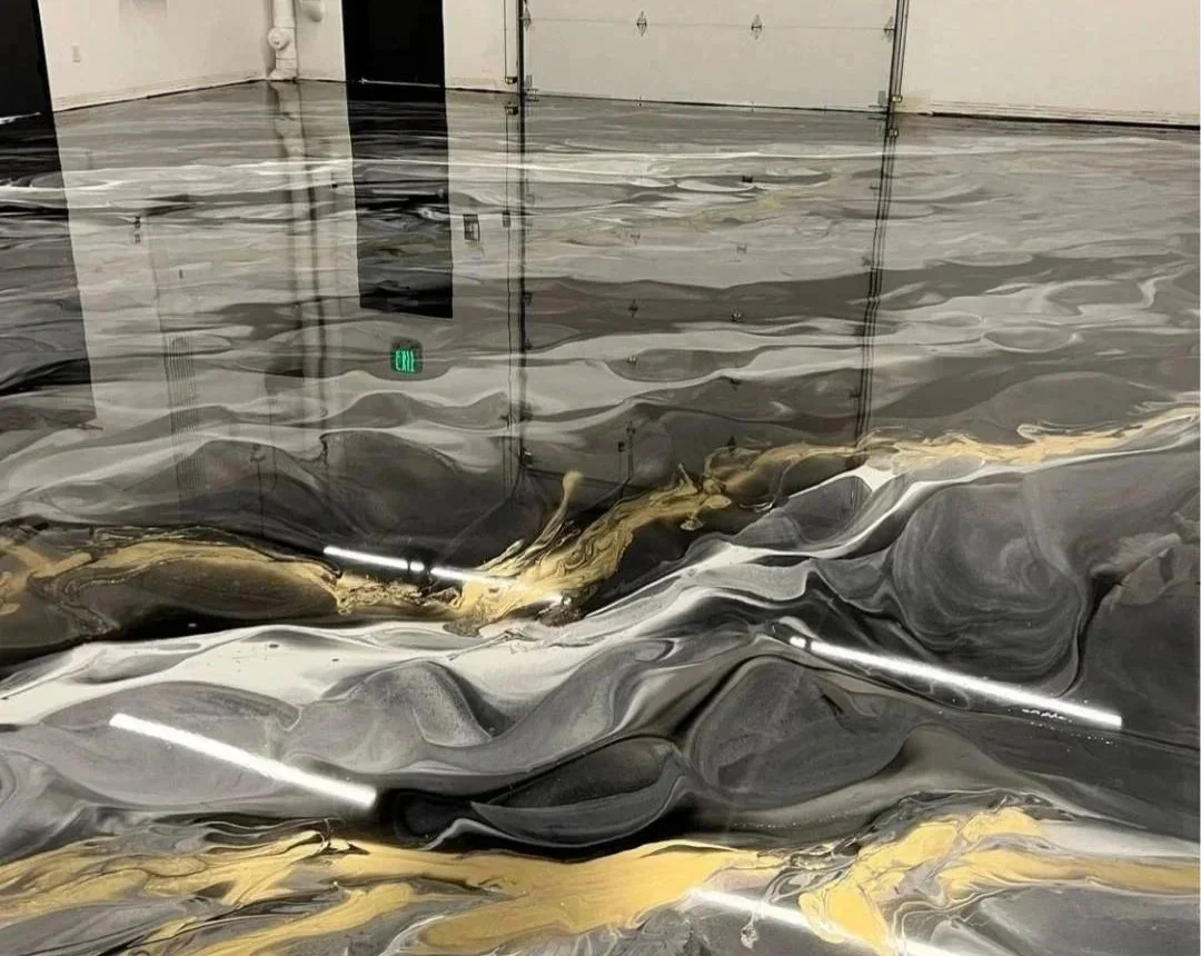 The image shows a highly reflective, marble-like epoxy floor with a swirling pattern of black, white, gold, and gray colors inside a building with a garage door and an exit sign reflected on the shiny surface.