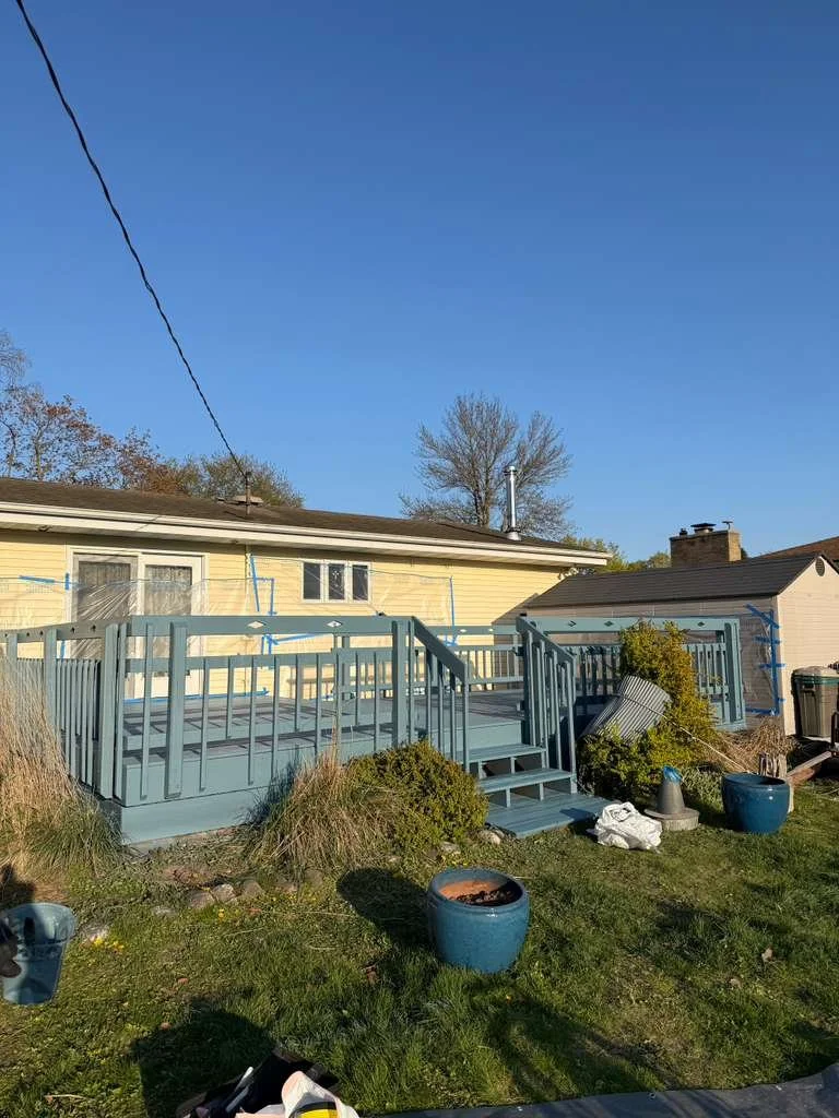 Backyard with a newly built light blue wooden deck, stairs leading down to grass, potted plants, and a yellow house with a brown roof in the background.
