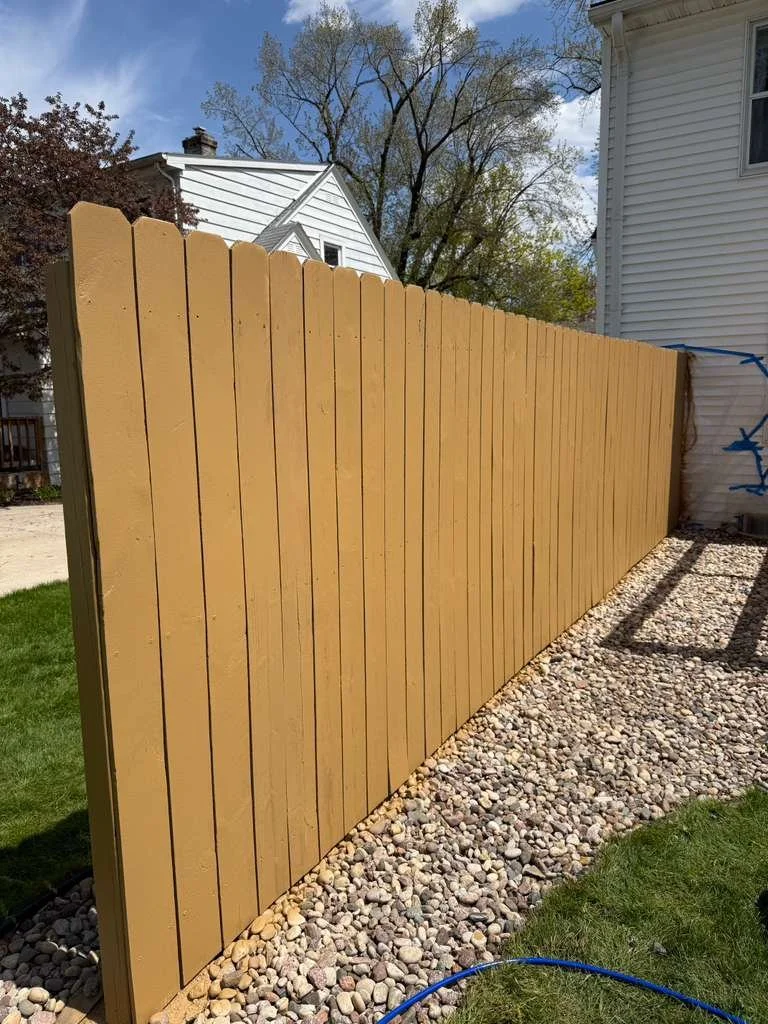 A newly built wooden privacy fence with vertical planks, painted tan, neighboring a white house, with a rock bed along the base and a grassy lawn adjacent.