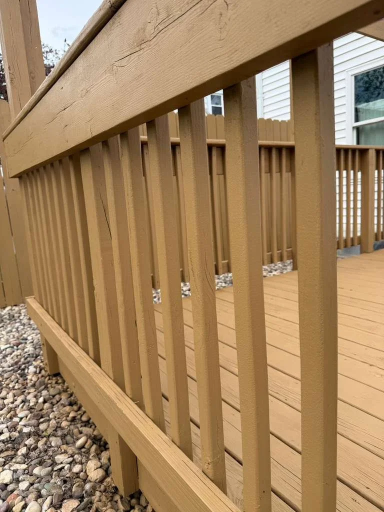 Close-up view of a newly built wooden deck with railing, showing vertical wooden balusters and a horizontal top rail, on a gravel ground.