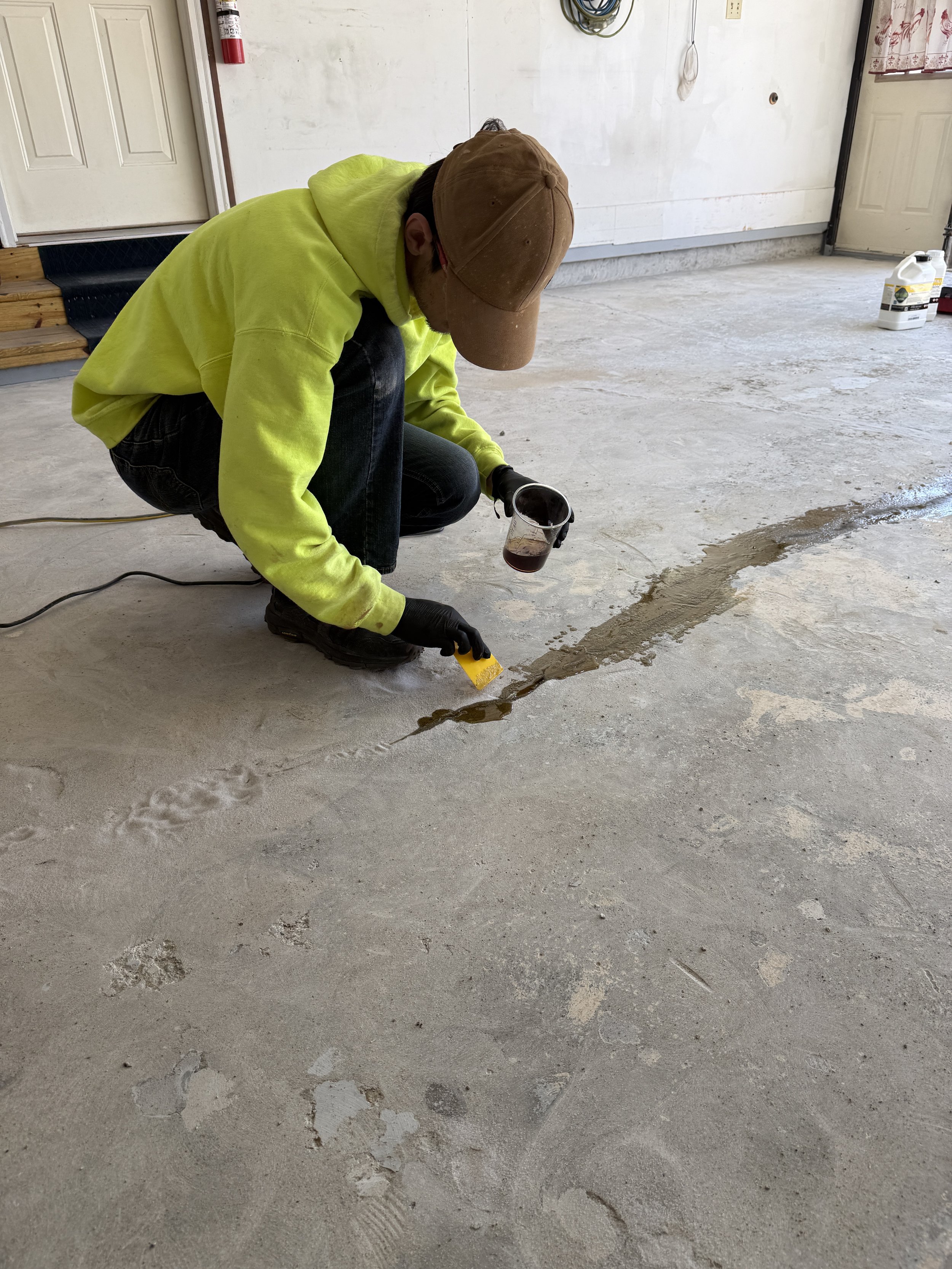 A person wearing a yellow hoodie, black gloves, black pants, and a brown cap is kneeling on the floor, applying a substance with a yellow trowel to a crack in a concrete floor. The person is holding a cup of dark liquid. The background shows a white 