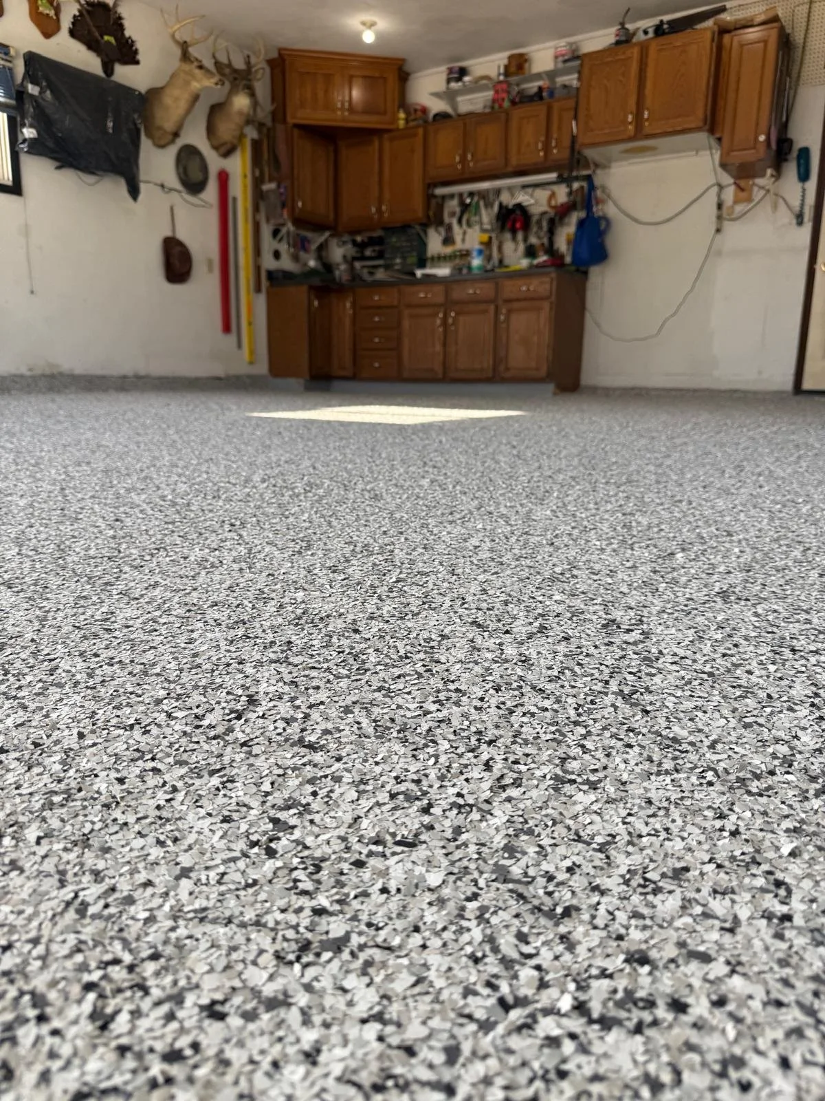 View of a garage interior with a gray speckled epoxy floor, wooden cabinets, and garage tools hanging on the wall. The garage features deer head mounts on the wall and various household items on shelves.