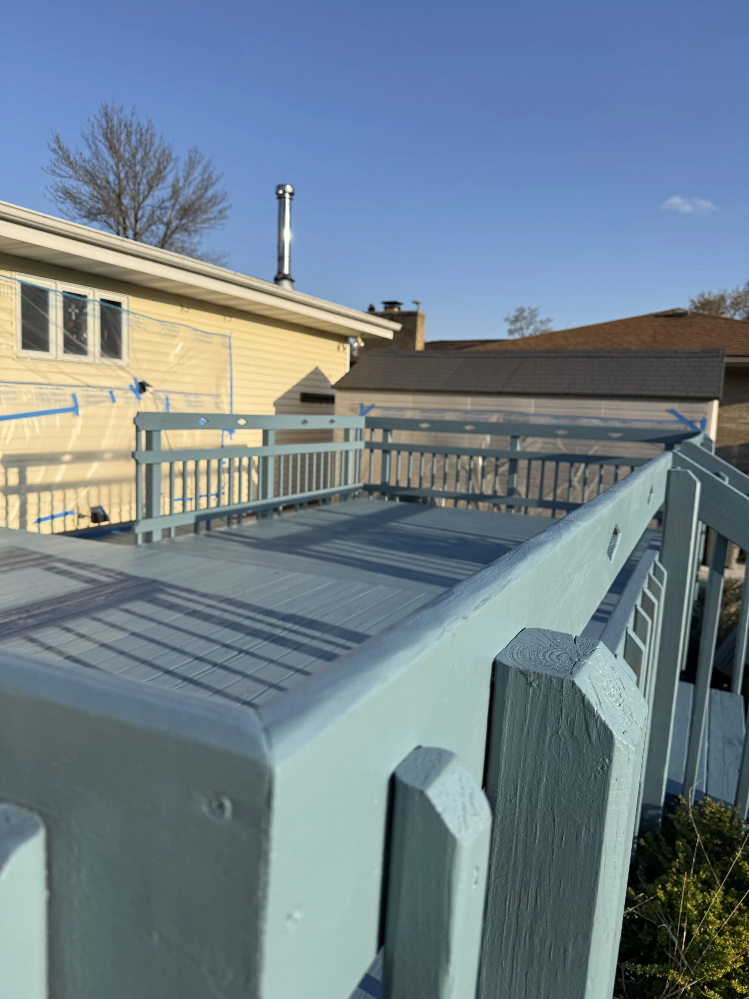 A light blue wooden deck with a railing, outside of a yellow house with a gabled roof, under a clear blue sky with a few clouds, and a large tree in the background.