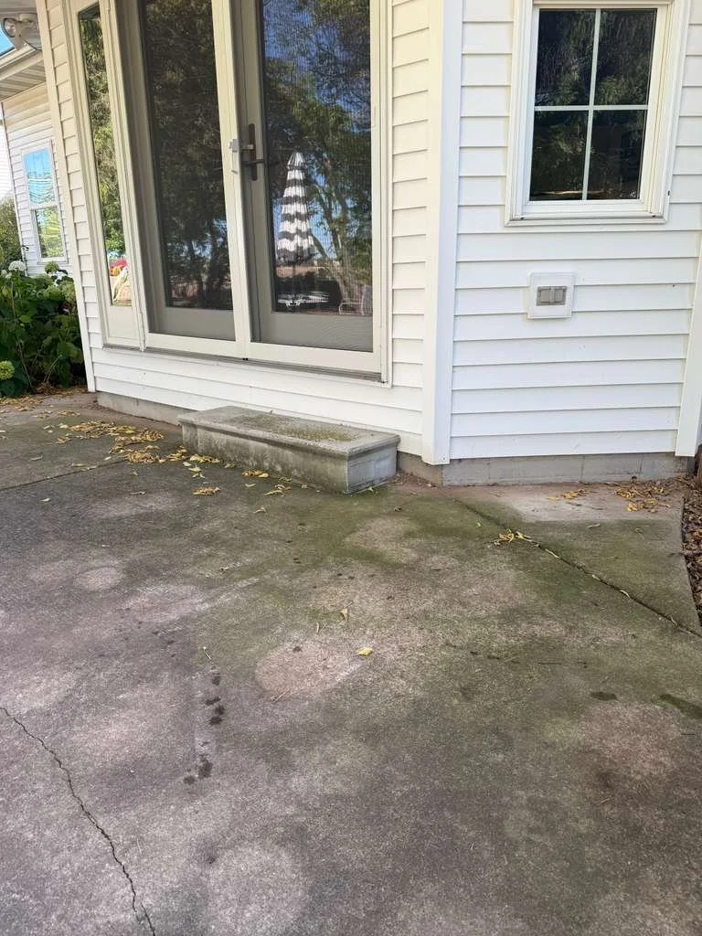 Backyard concrete patio with a small step, adjacent to a white house with vinyl siding, a sliding glass door, and a window. There are some fallen leaves on the surface.