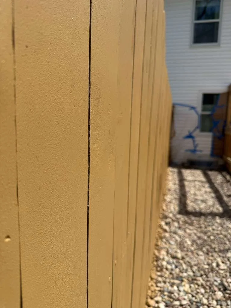 Close-up of a beige wooden fence with a gravel ground behind it, and a house with white siding and windows in the background.