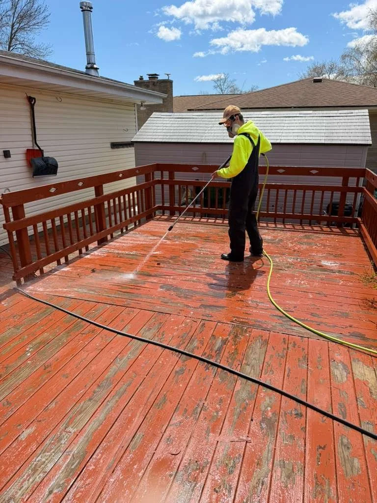 Person wearing a yellow and black hoodie, black pants, sunglasses, and a cap power washing a wooden deck on a sunny day.