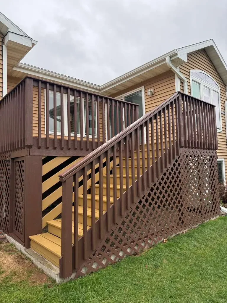 A residential exterior with a wooden deck staircase leading up to a sliding glass door and a window on a beige house with brown trim, led by brown railings and lattice skirting, on a grassy lawn.