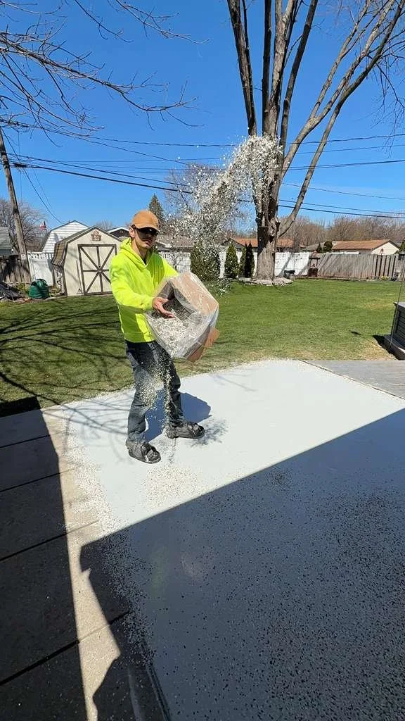 A person wearing a neon yellow jacket, sunglasses, and a cap is throwing white salt or sand onto a concrete patio in a backyard, likely for ice melting or snow removal, with clear blue skies and a tree in the background.