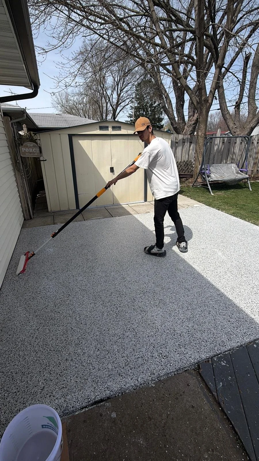 A young man wearing a tan cap, white t-shirt, black pants, and roller skates is using a long-handled roller to apply a speckled coating on a small backyard patio. The patio is adjacent to a house with beige siding and a white shed with black trim. In