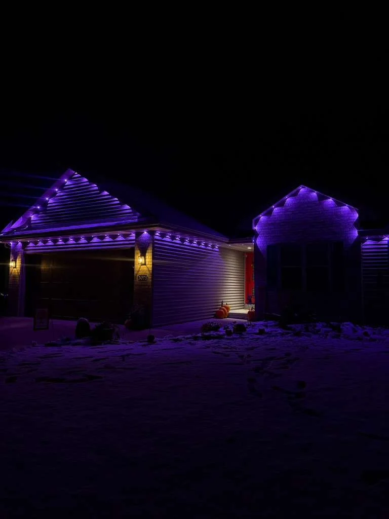 A house decorated with purple Christmas lights outlining the roof and gables at night, with a ghost decoration illuminated on the left side of the yard.