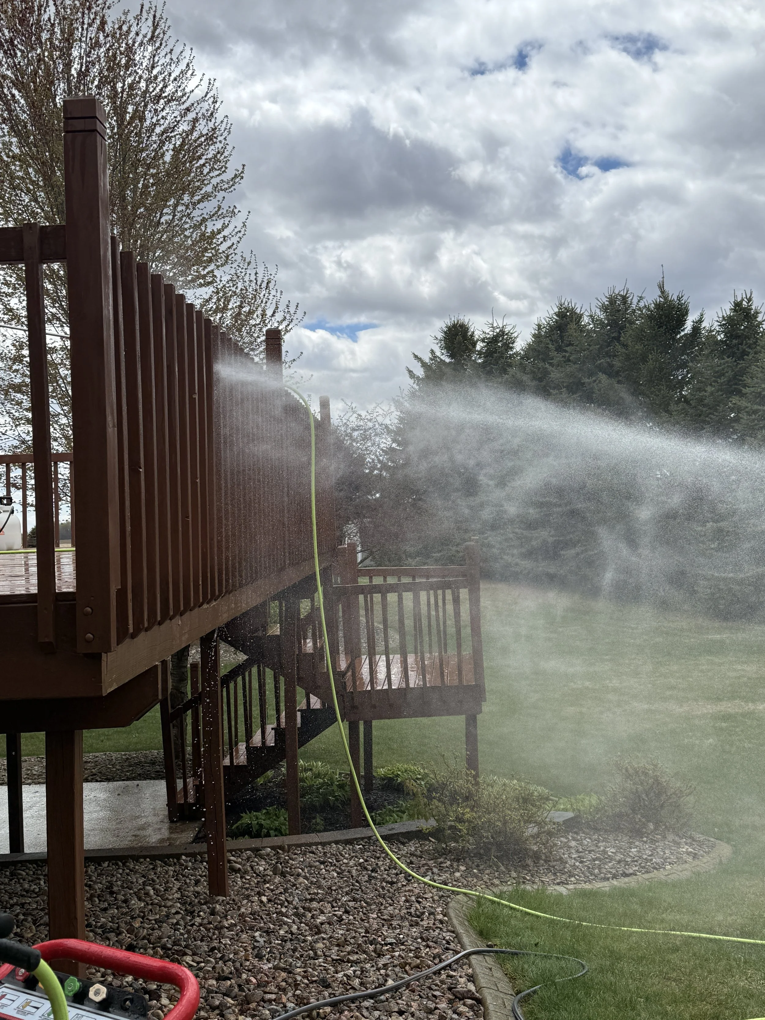 A backyard scene with a wooden deck and a hose spraying water, creating a mist. In the background, there are trees and a cloudy sky.