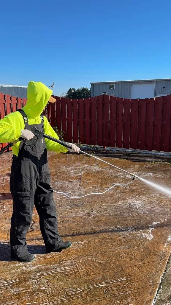 Person wearing a neon yellow hoodie, black overalls, and white gloves power washing a wooden deck in a backyard with a red fence and blue sky.